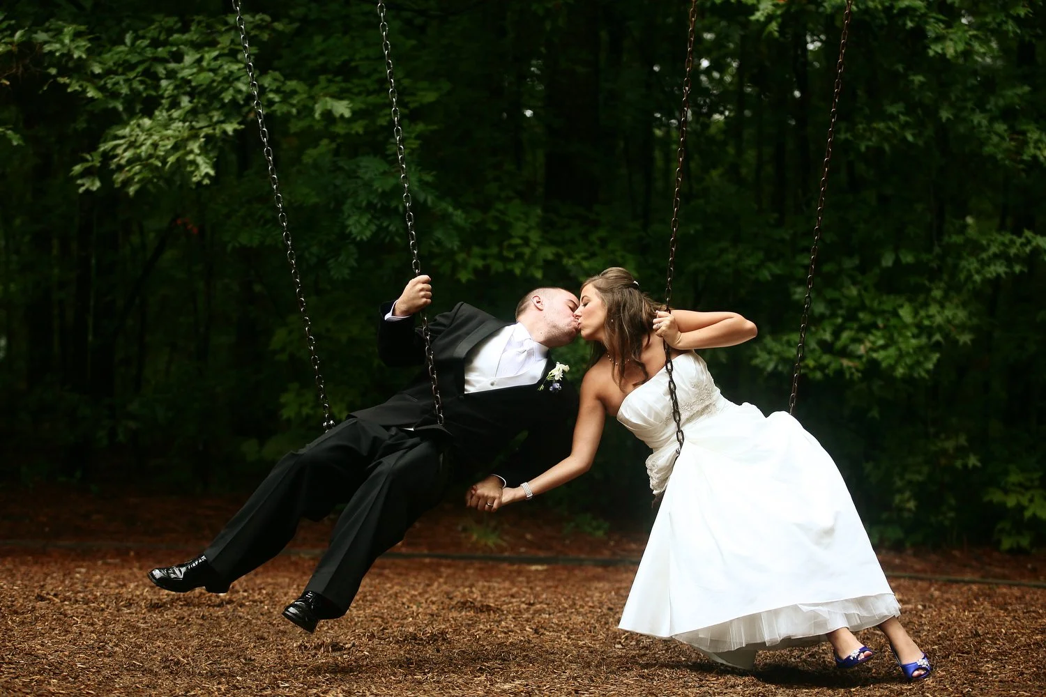 A bride and groom in wedding attire sitting on swings outdoors, lean in to kiss each other amidst a background of dark green trees. The groom is wearing a black suit. The bride has a strapless wedding dress and dark blue satin shoes.