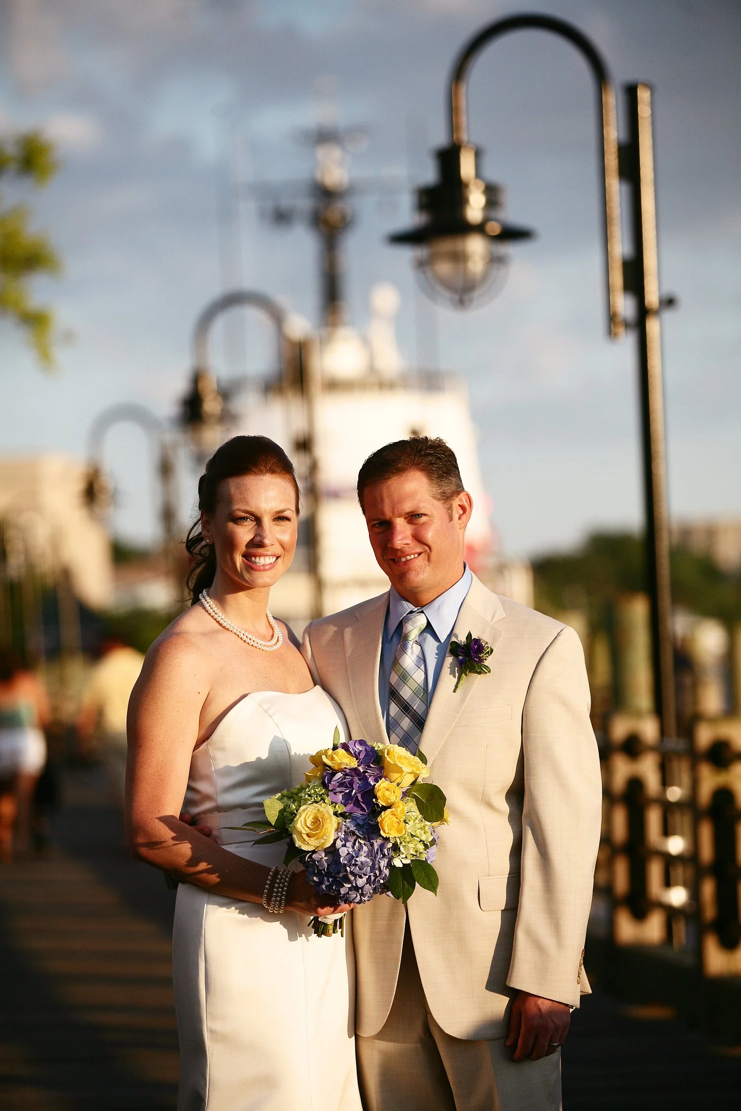 A bride and groom posing outdoors on a wedding day, with lampposts and a boat in the background, during sunset.