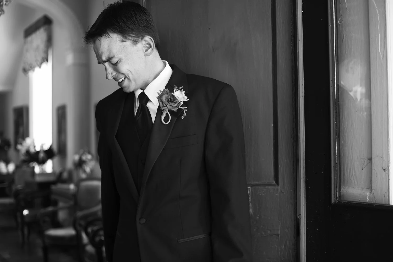 A young man in a suit with a boutonniere, with an emotional expression on his face as he leans against an open doorway with natural light coming into the room.