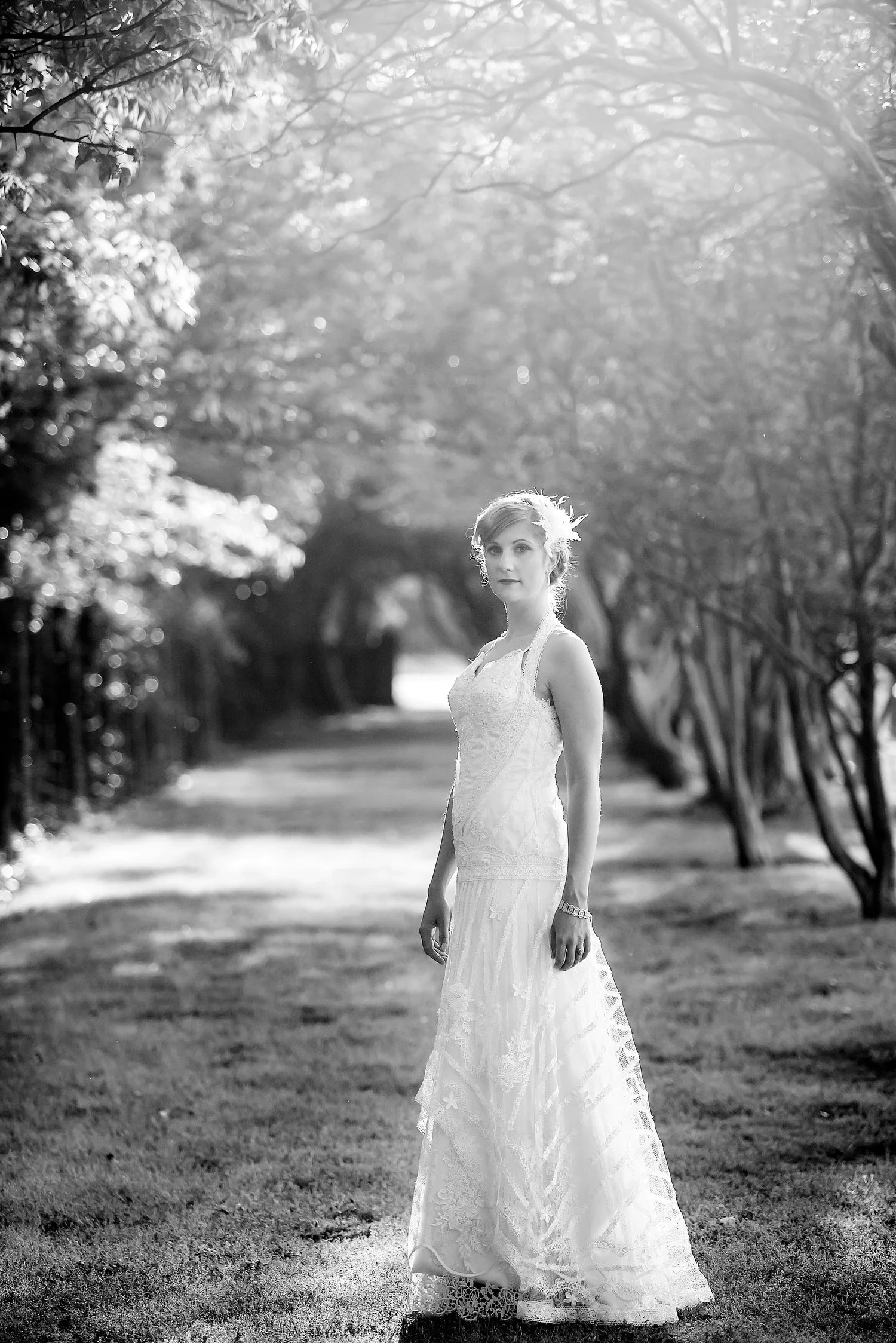A woman in a wedding dress standing on a path in a wooded area, illuminated by sunlight filtering through trees.