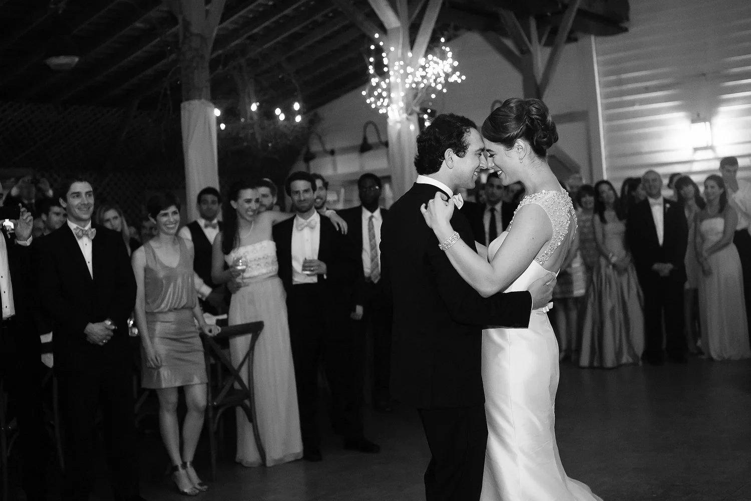 Black and white photo of a newly married couple dancing at their wedding reception, surrounded by friends and family in an indoor venue with hanging lights and wooden beams.