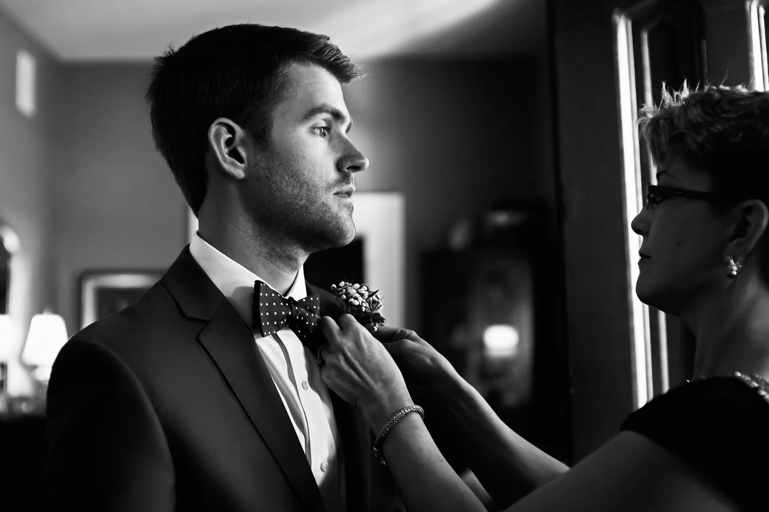 Black and white image of a man in a suit and bow tie looking serious as a woman adjusts his bow boutonniere.