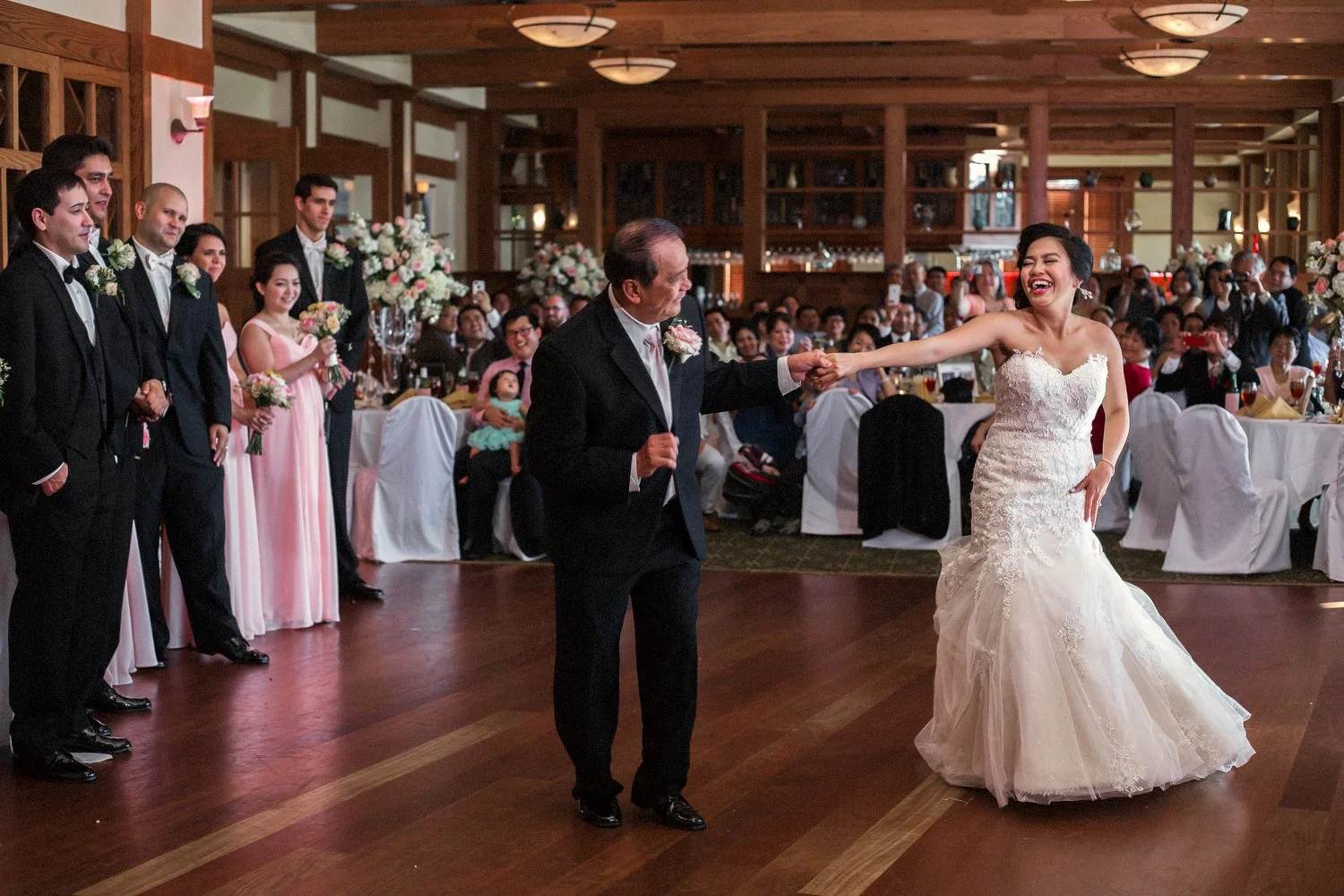 A wedding reception with wooden floors and wooden decorative walls. The wedding party, men in black suits and women in long pink dresses, line one side of the dance floor. Guests are seated at tables in the background. A bride in a strapless white we