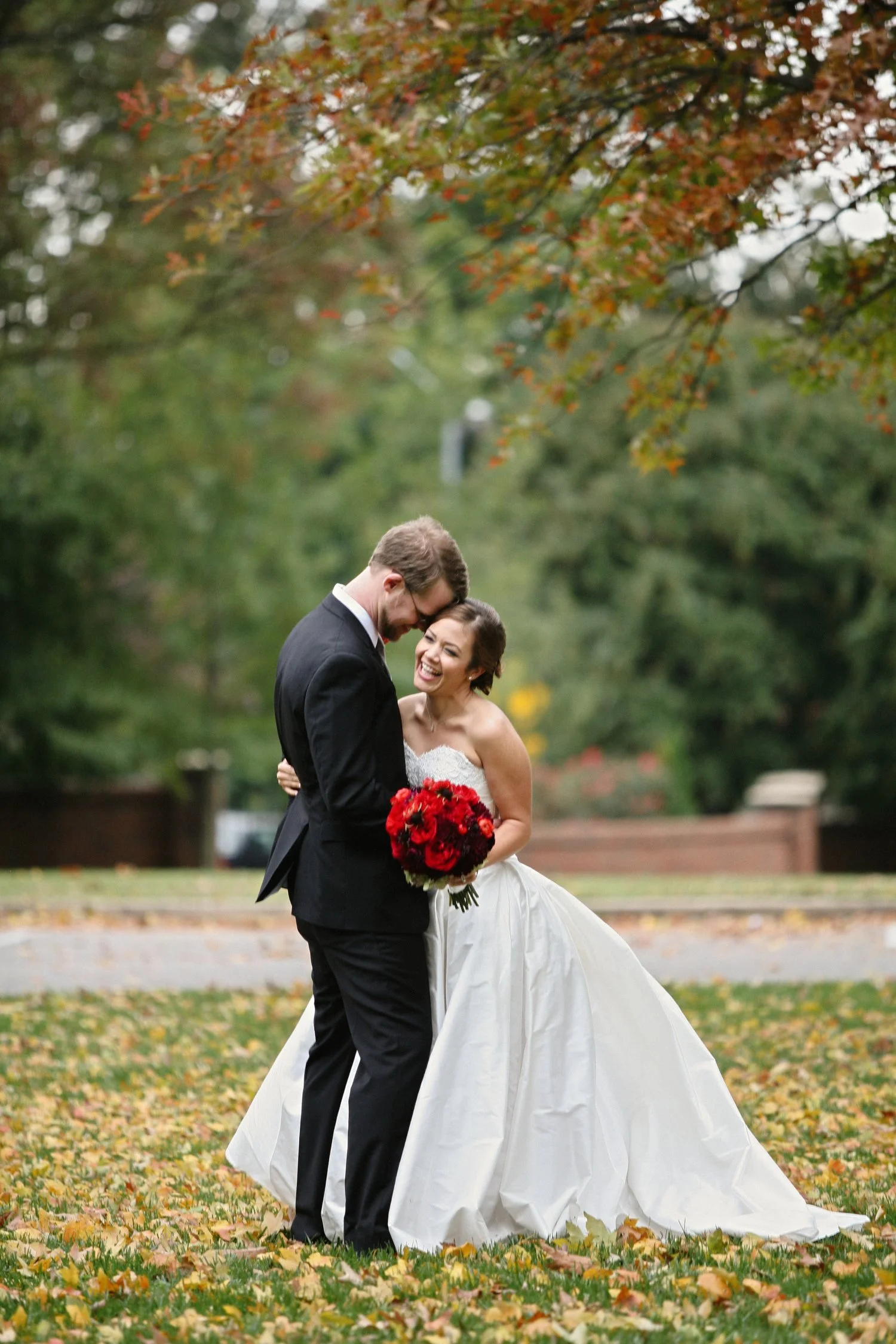 A newlywed couple dressed in wedding attire standing on a grassy area with fallen autumn leaves, embracing and smiling, surrounded by trees with fall foliage. She is holding a bouquet of red roses.