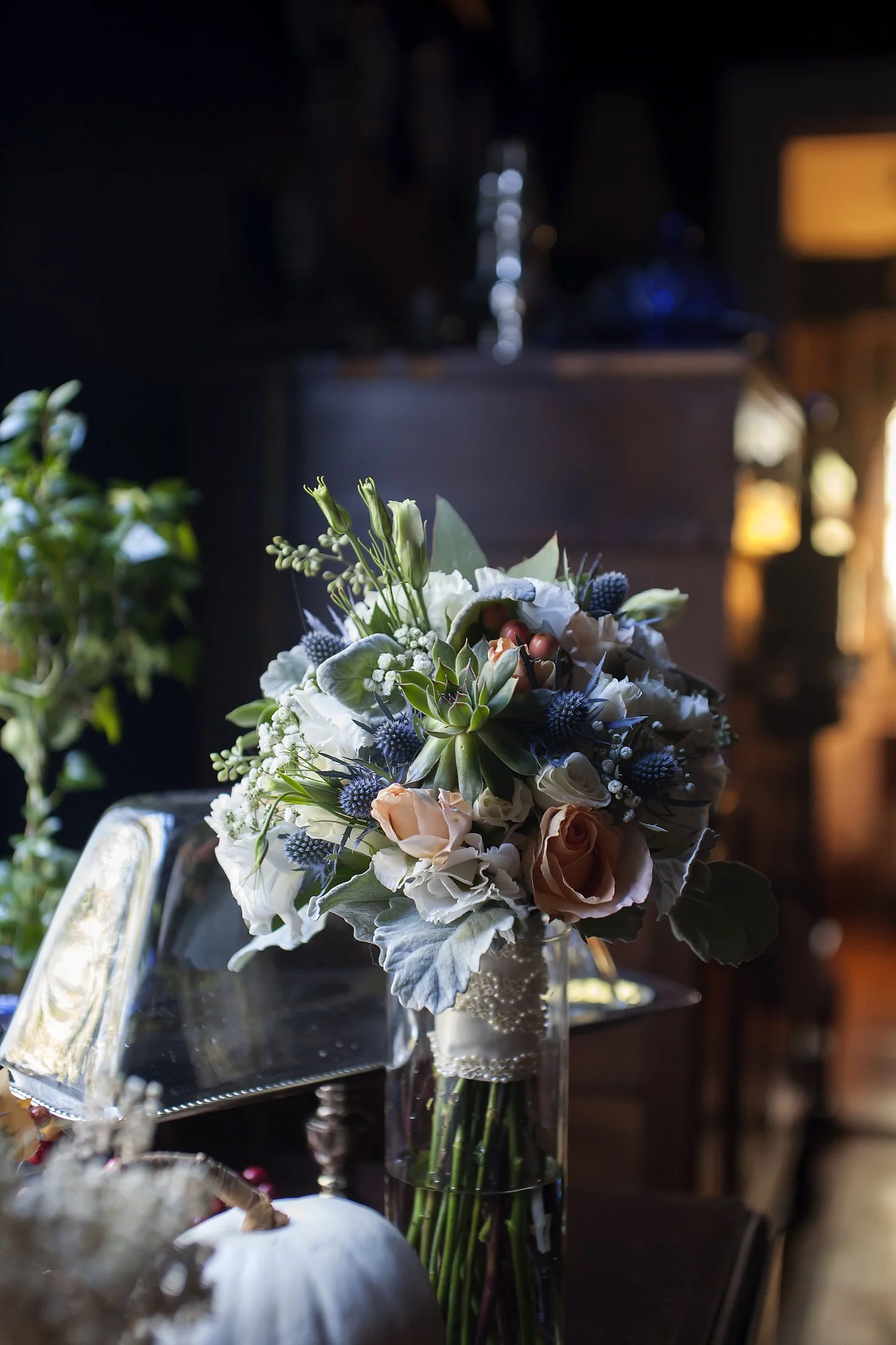 A bridal bouquet of peach, blue and white flowers wrapped in lace sits in a glass jar of water on a wooden table in window light. Other items are around including a small white pumpkin, a vintage lamp and a green plant.