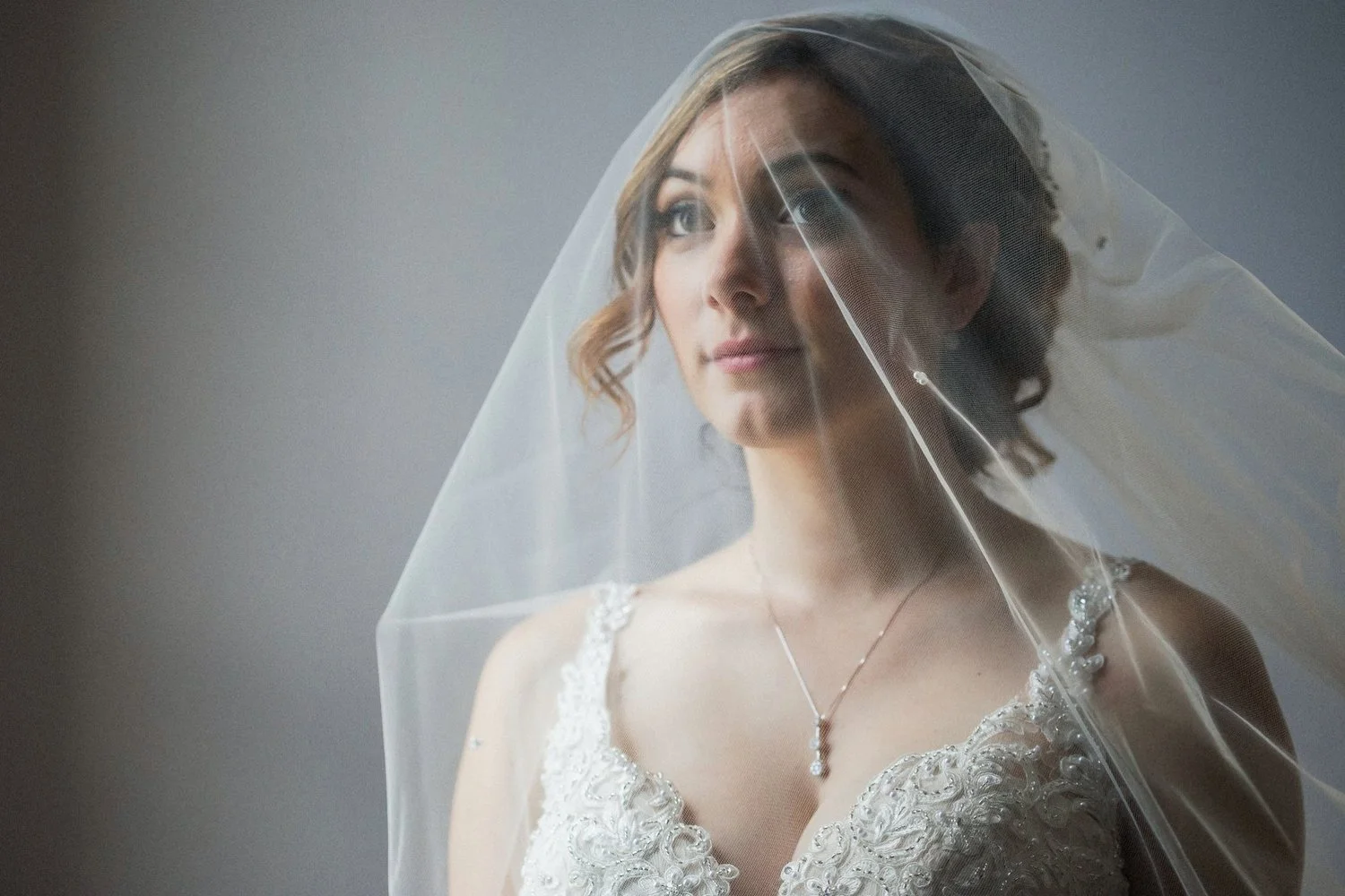 A bride in a white lace wedding dress and veil over her face gazing thoughtfully upward.