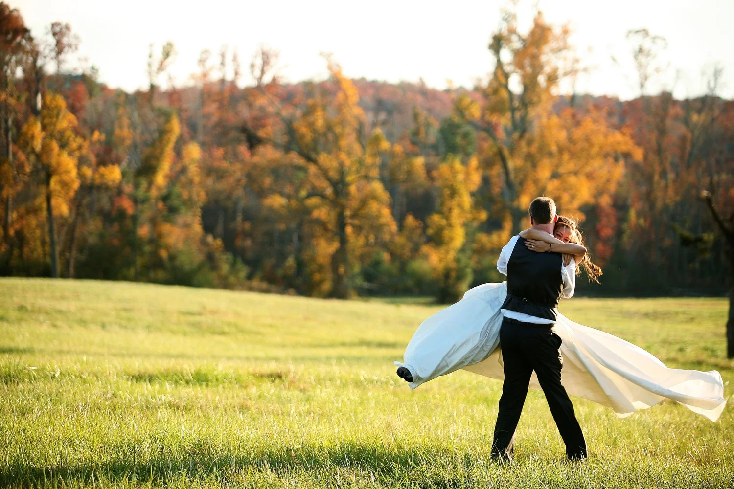 A groom holding a bride in a white wedding dress in a grassy field with autumn trees in the background. He is swinging her in the air and her dress is flowing around her. Her long reddish-brown hair is long and in loose curls blowing to the side as h