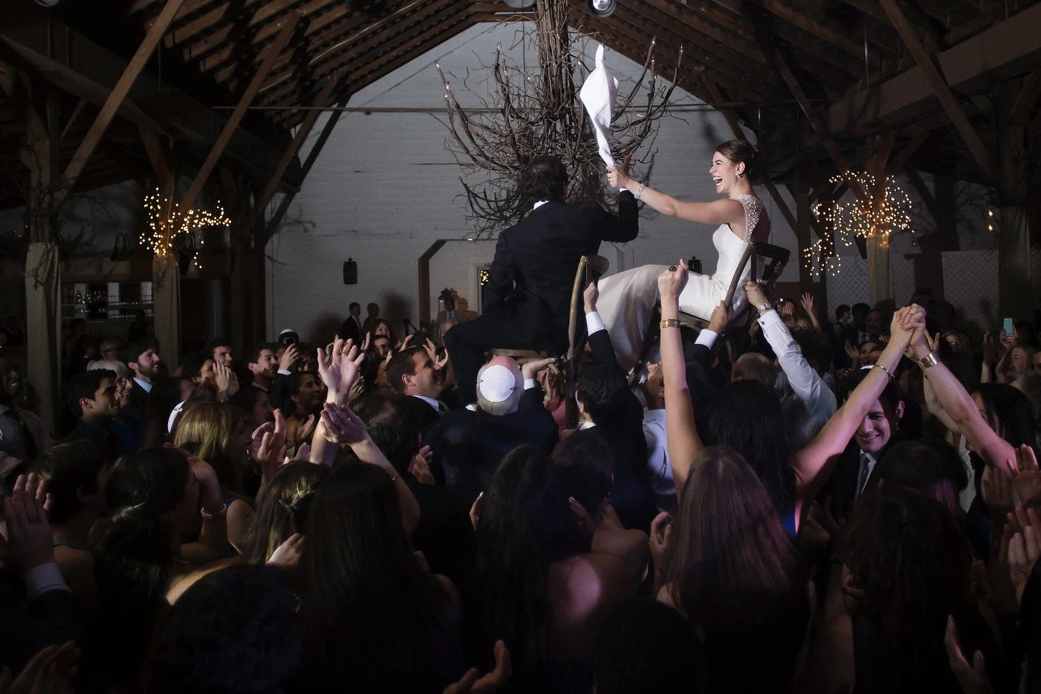A bride and groom are being held up above the dance floor in chairs by their guests. She is seen from the side wearing a white wedding dress and holding something white (perhaps a napkin) up in the air with her left arm as she grins. The groom holds 
