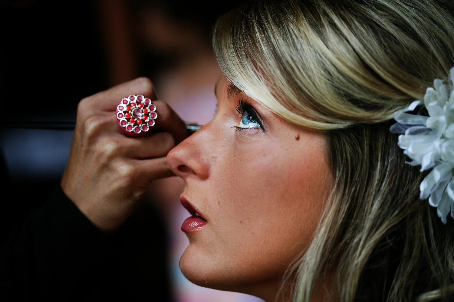 A woman getting her makeup done, with focus on her face as someone applies makeup near her nose. The woman has long blond hair with a white flower bow in it. The hand applying her makeup has a large round ring with bright pink beads on it.