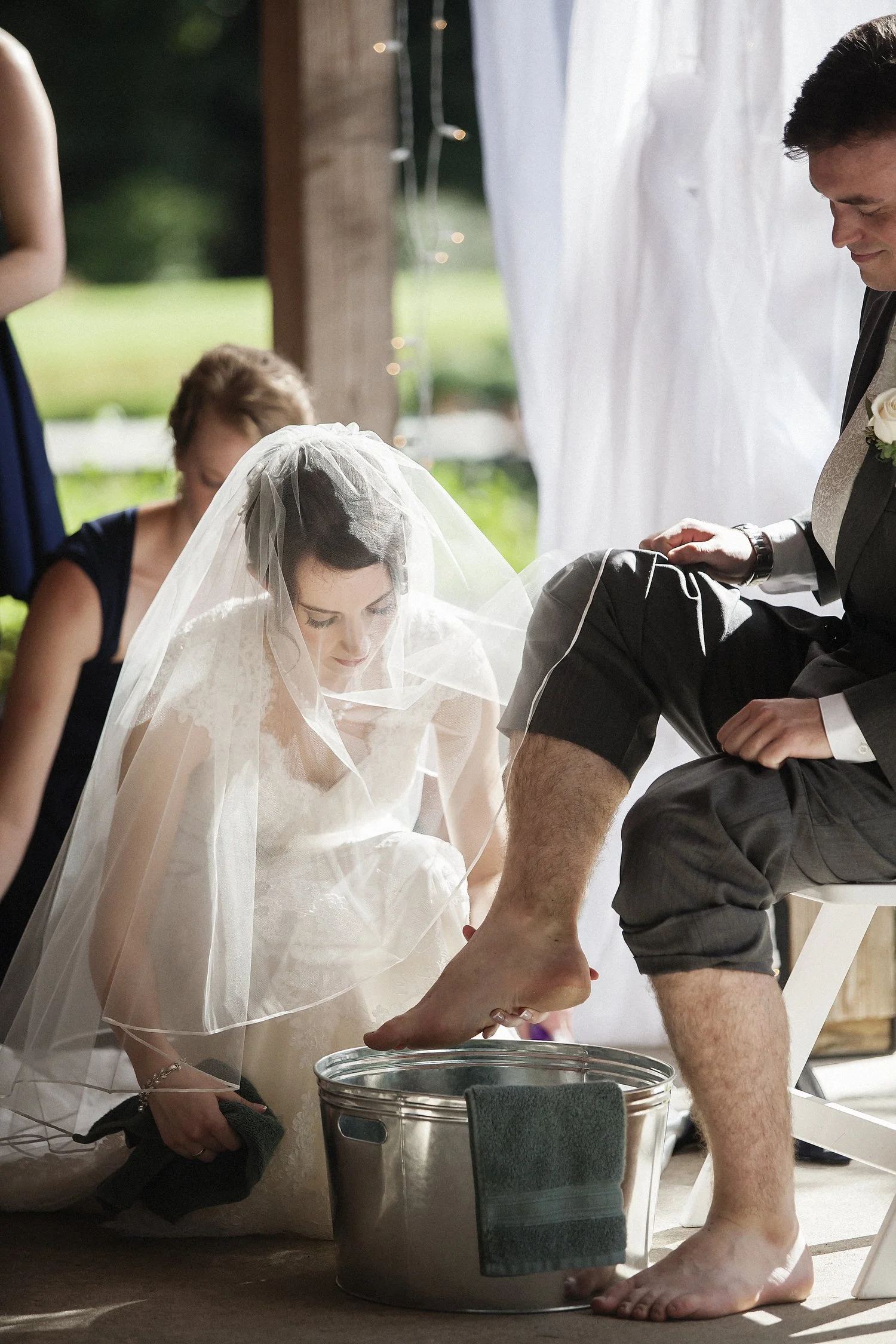 A bride with a veil washing her groom's feet in a silver basin as part of their outdoor wedding ceremony under a wooden structure with sheer fabric hanging.