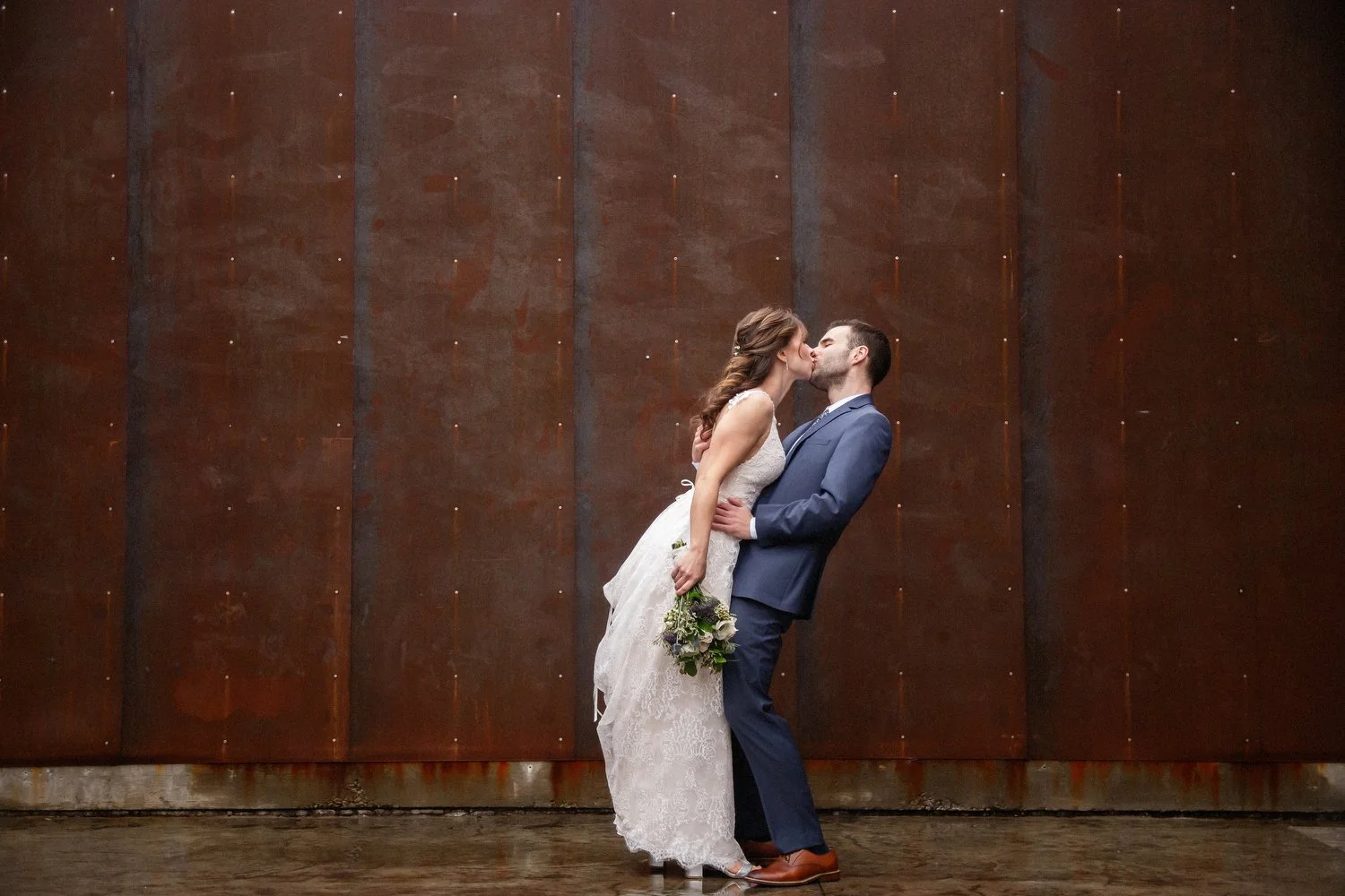 A bride leans over to kiss her groom as they stand closely in front of a dark brown metal wall, the bride holding a bouquet of flowers, with the groom wearing a blue suit.