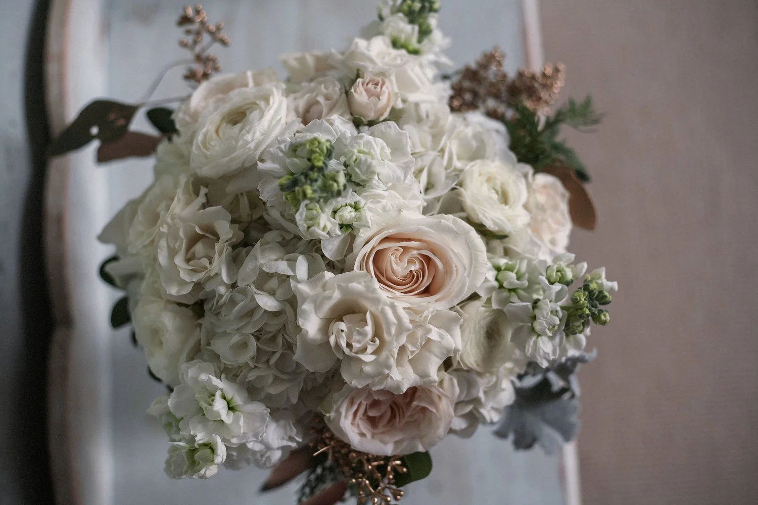 Close up image of a bridal bouquet with pale-colored flowers. There are pink roses, white ranunculus and other smaller pink and white flowers. Lamb's-ear leaves hang around the bottom. 