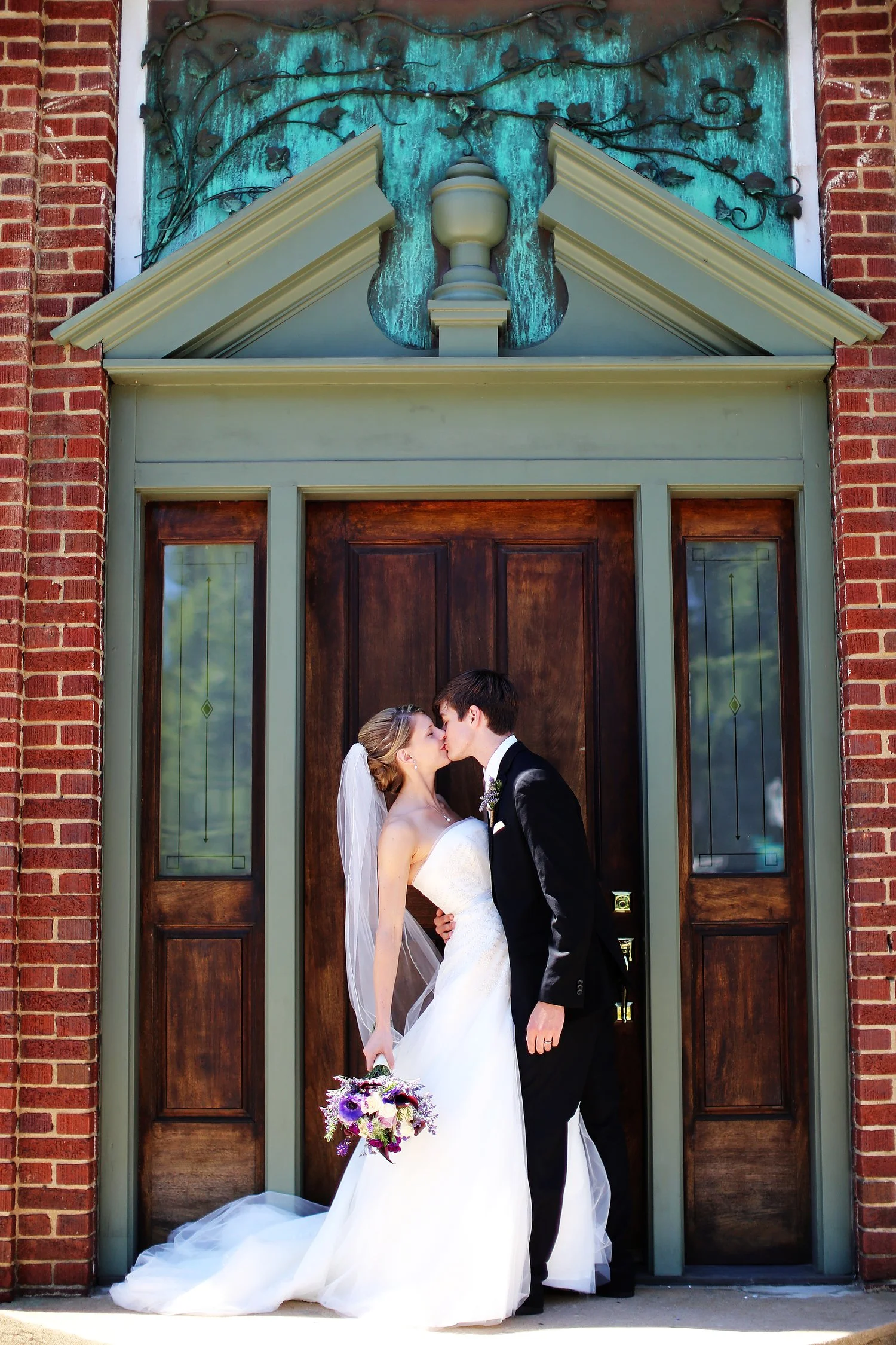 A bride and groom kiss outside a dark wooden door with brick walls and decorative stained glass all around. She is wearing a strapless white wedding dress and a white veil and clutching a bouquet of flowers at her side. He has on a black suit.