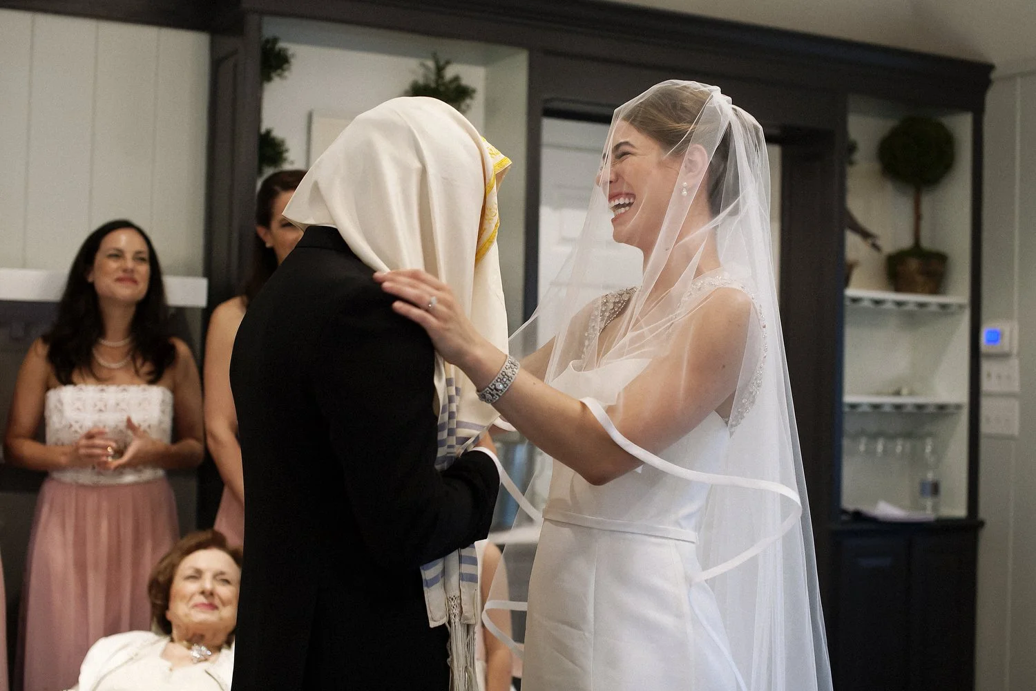 Bride and groom at their Jewish wedding ceremony, smiling and holding each other, with guests observing in the background. She has on a white dress and a sheer veil over her face. He has on a black suit and a special cloth is wrapped around his head.