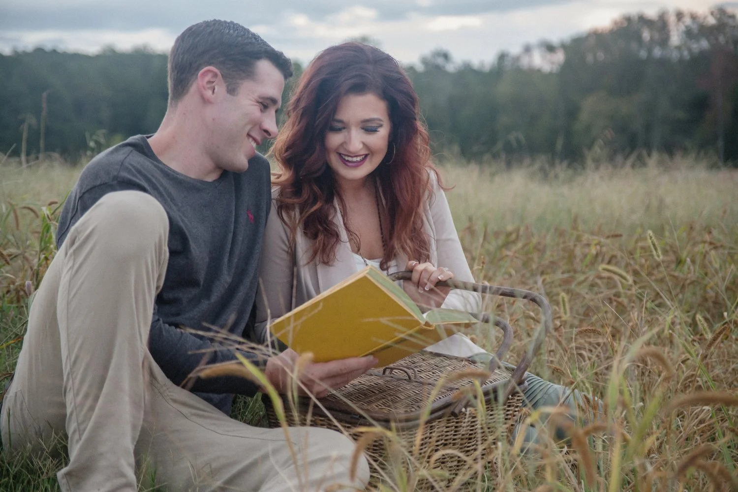 A couple sits together in an overgrown golden grassy field with dark green trees in the distance behind them. They are holding a book with a yellow cover and smiling as they look at it together. He has on a grey long-sleeved shirt and khakis. He sits