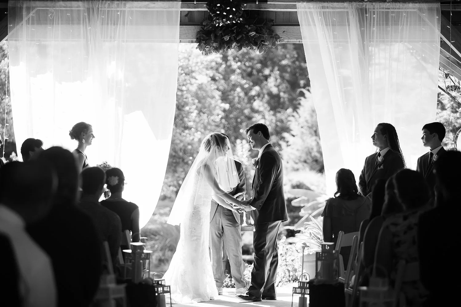 A black and white photo of a wedding ceremony with a bride and groom holding hands and exchanging vows, standing beneath a canopy with curtains and hanging decorations. Guests are seated on either side, and bridesmaids and groomsmen stand nearby.