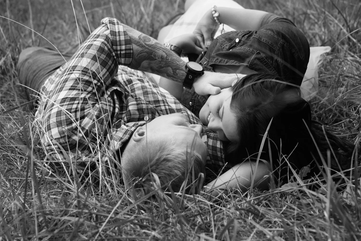 Black and white photo taken from above of a couple lying down in an overgrown grassy field. They are turned facing each other about to kiss. He has on a flannel and jeans. She has on a dark colored shirt or dress.