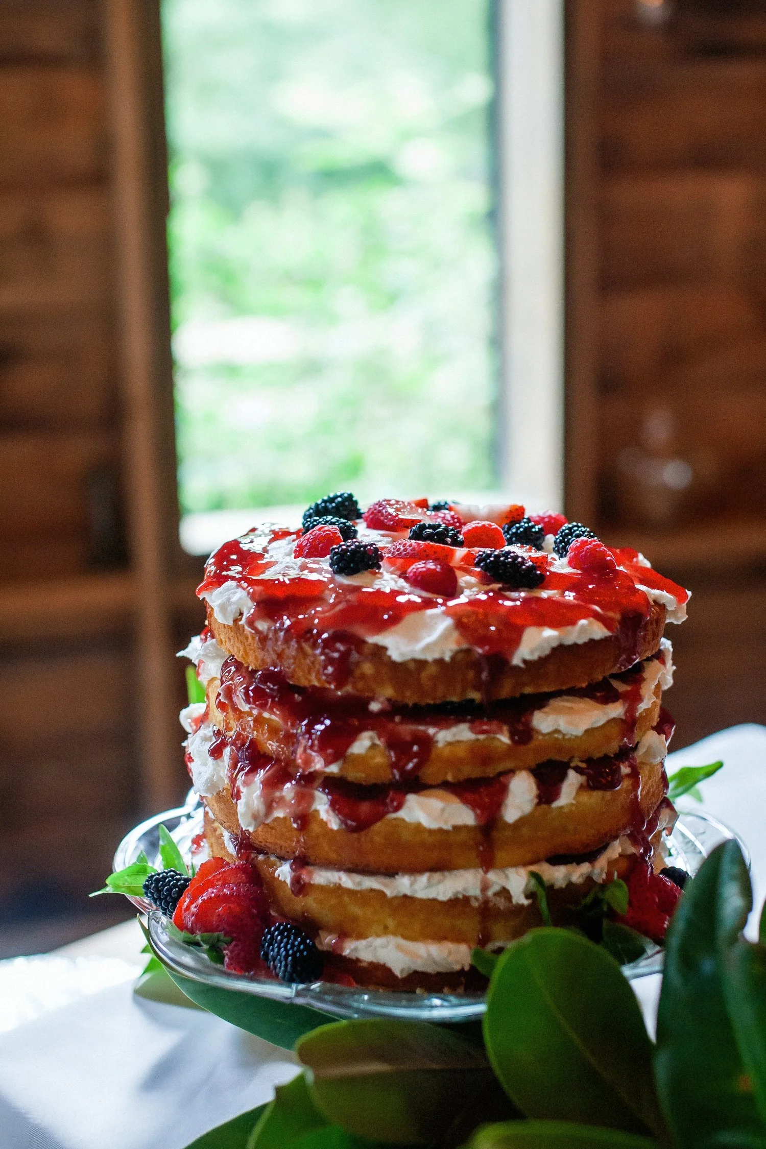 Five-tiered naked cake is adorned with cream cheese frosting and strawberry jam between layers. On top are berries and more cream cheese frosting. An open-air window is behind the cake and green trees are far in the distance. The walls around the win