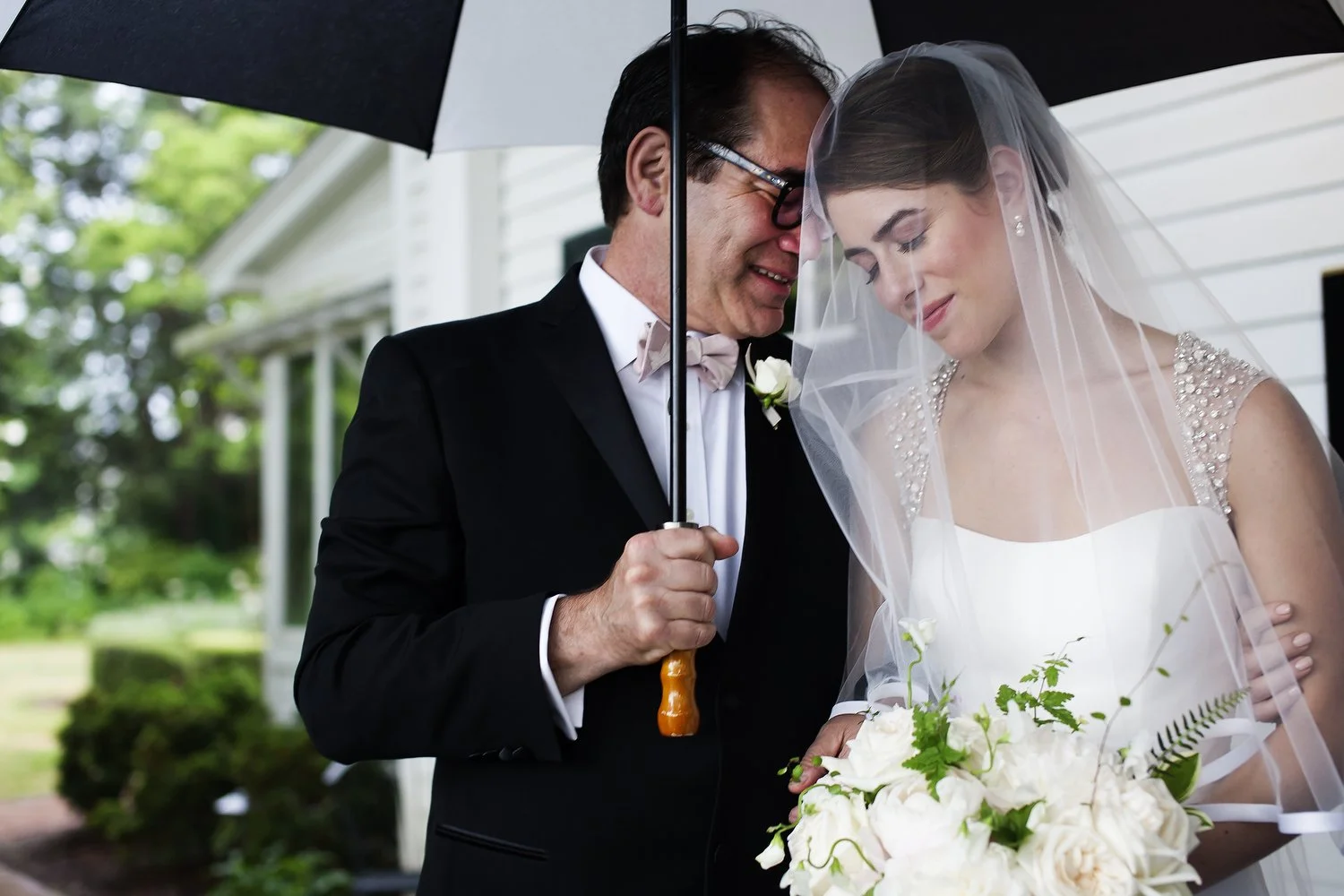 A bride and an older man, possibly her father, sharing a tender moment under an umbrella on her wedding day. The bride wears a white wedding dress with beaded details and a veil, holding a bouquet of white roses and greenery. The man is in a black tu