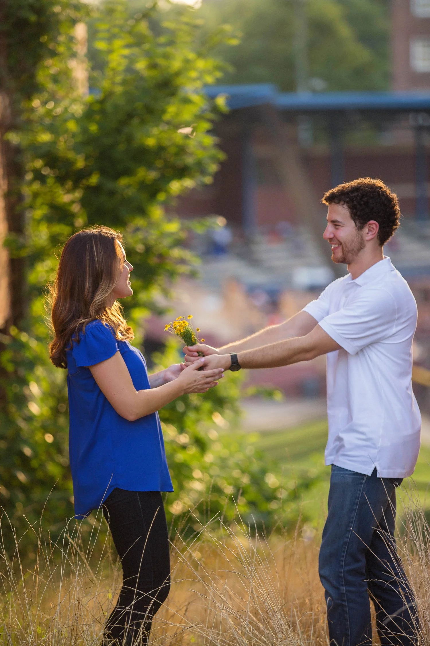 Vertical photo of a couple standing facing each other and holding out their arms to hold hands. She is wearing a blue shirt and black leggings. He has on a white shirt and jeans. They are in a golden field and there is an old building and a bright gr