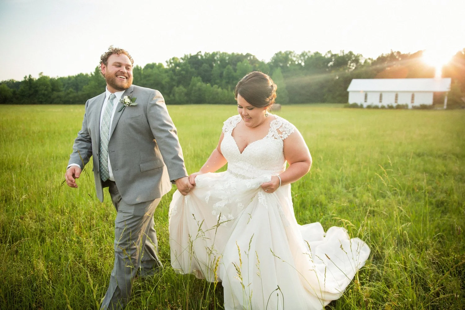 A small white church is seen in the far right corner in the distance. A bride and groom hold hands and walk through a lush green field in the foreground. He has on a grey suit. She has a white v-neck lacy wedding dress with cap sleeves. He helps her 