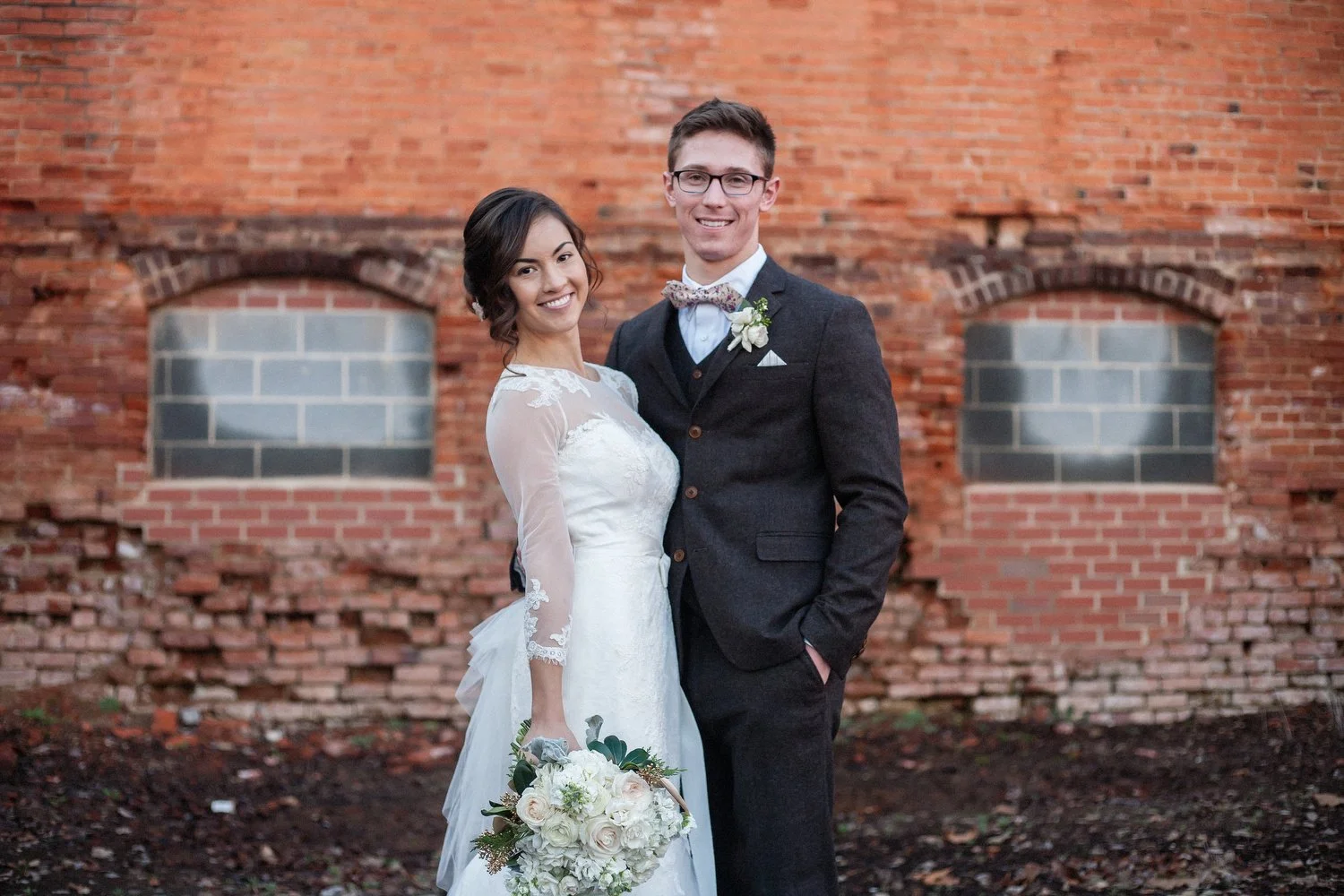 A bride and groom in wedding attire standing close together outdoors, smiling with a weathered brick wall behind them.