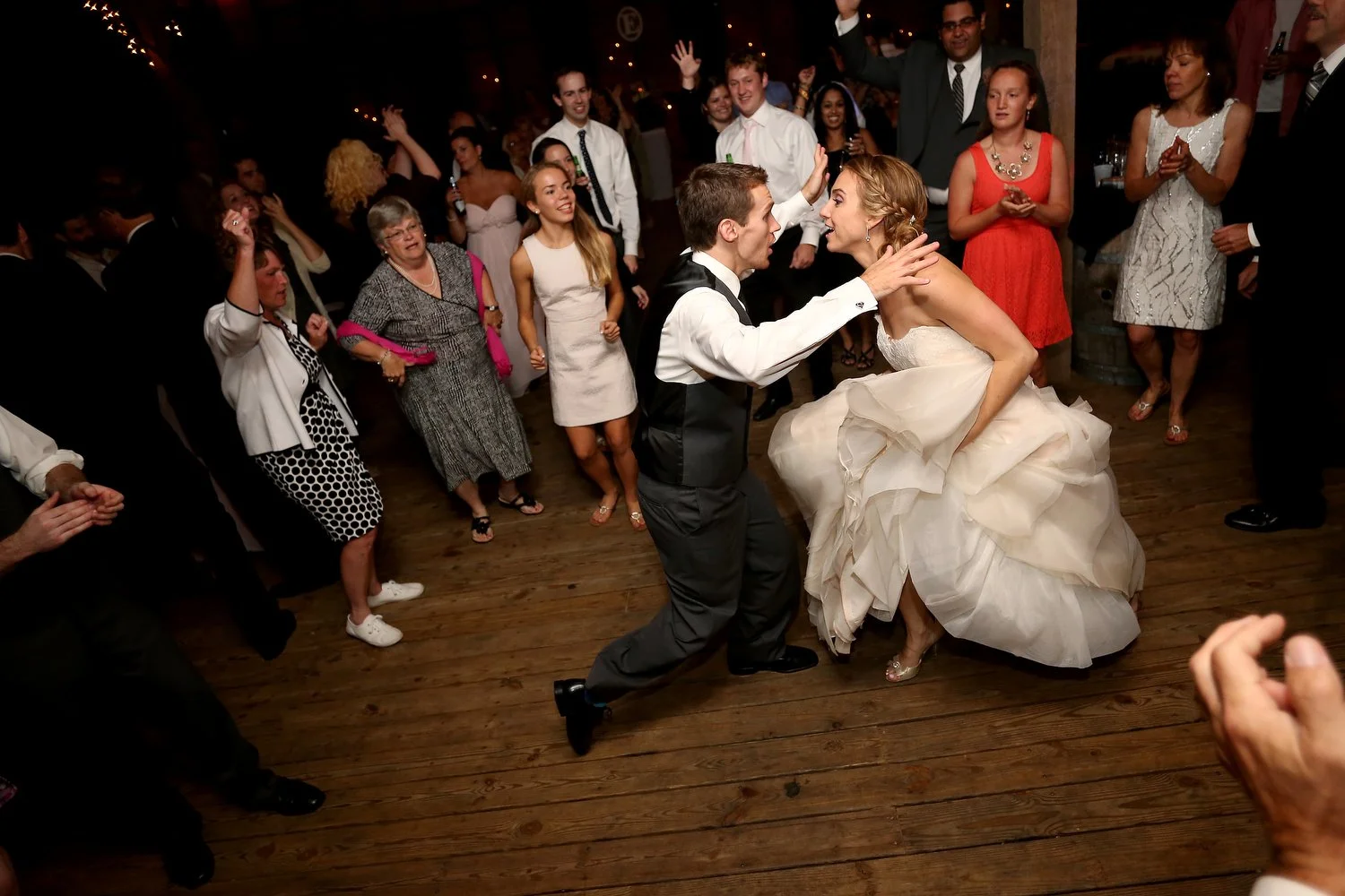 A bride and groom kneel in the center of the dance floor as guests surround them watching and dancing. The groom has on dress pants and a vest with a white dress shirt. He is kneeling and has his right arm up on her shoulder. She has on a white strap