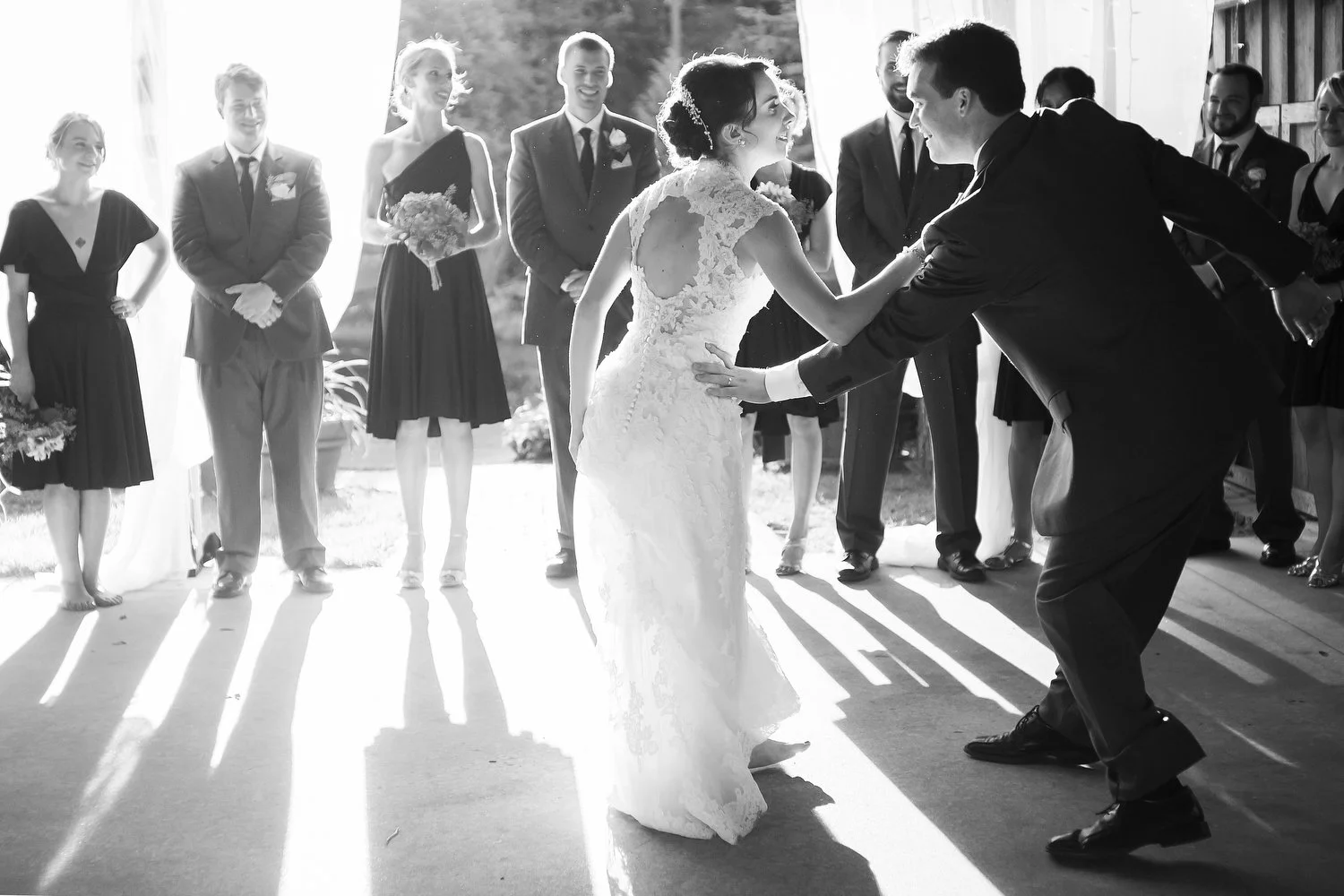 Black and white photo of a wedding reception with bride and groom dancing, surrounded by wedding party and guests in formal attire, indoors with sunlight streaming through windows.