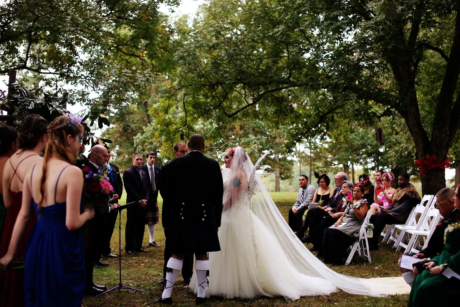 A wedding ceremony outdoors under a large tree with draped leaves. The bride in a white gown with a train and veil faces a groom, who wears a traditional Scottish kilt. Bridesmaids in colorful dresses and groomsmen in suits stand nearby, and guests s