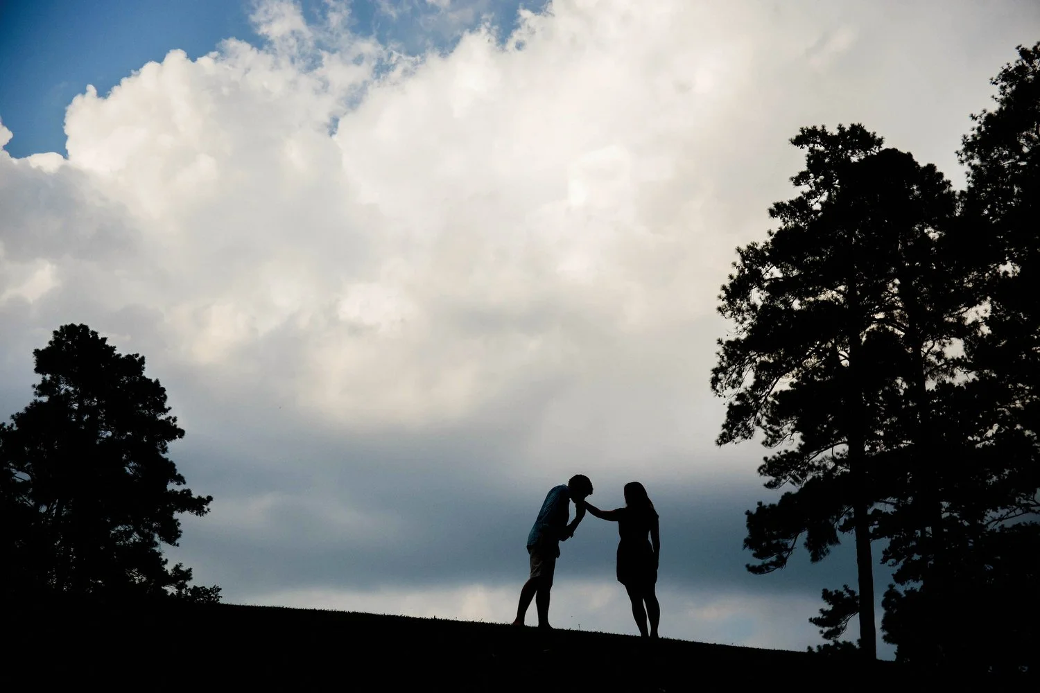 Silhouette of a couple in a field with tall pine trees to their right and left and huge white puffy clouds filling the sky behind them. The couple is small in the frame. She is standing facing him and holding out one arm. He leans in from a distance 