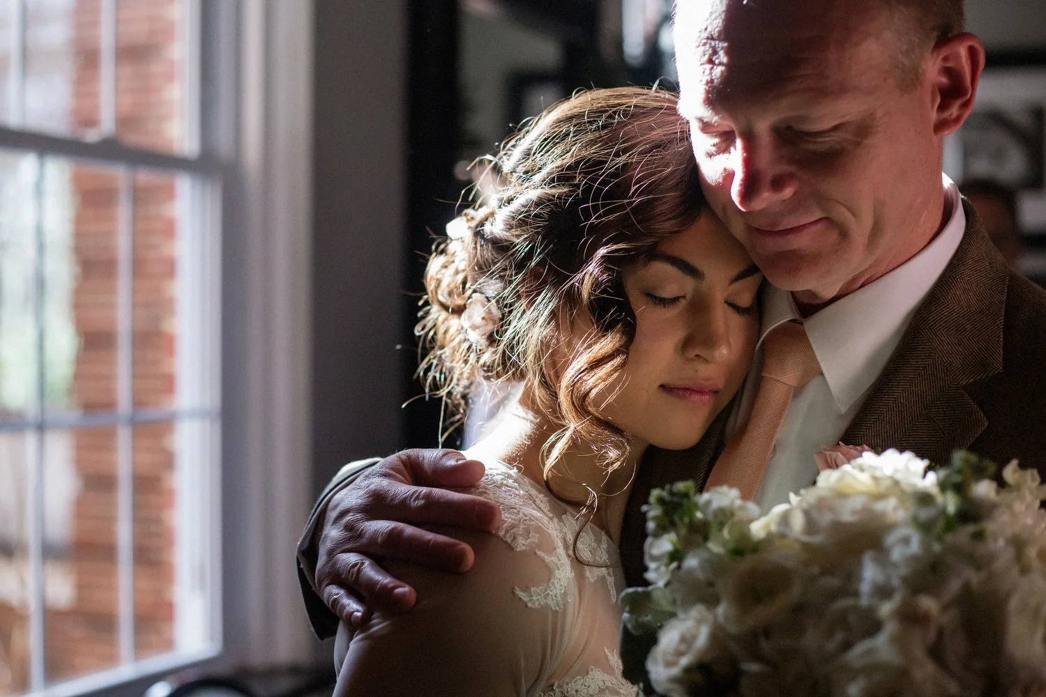 A bride in a lacy long-sleeved wedding dress rests her head on her father's shoulder by a window with natural light before he walks her down the aisle. She is holding a bouquet of white flowers.
