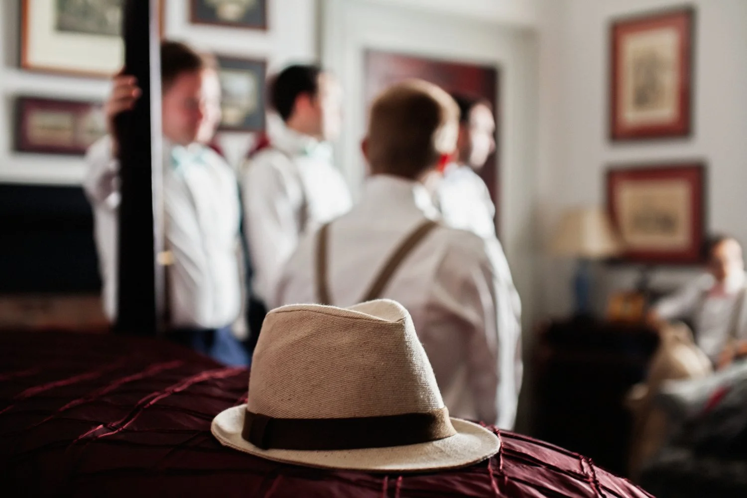A tan fedora sits in the foreground on the corner of a bed with a dark rust-colored bedspread and brown wooden post. In the distance men with white dress shirts and suspenders are in a group talking with each other. Various picture frames are on the 