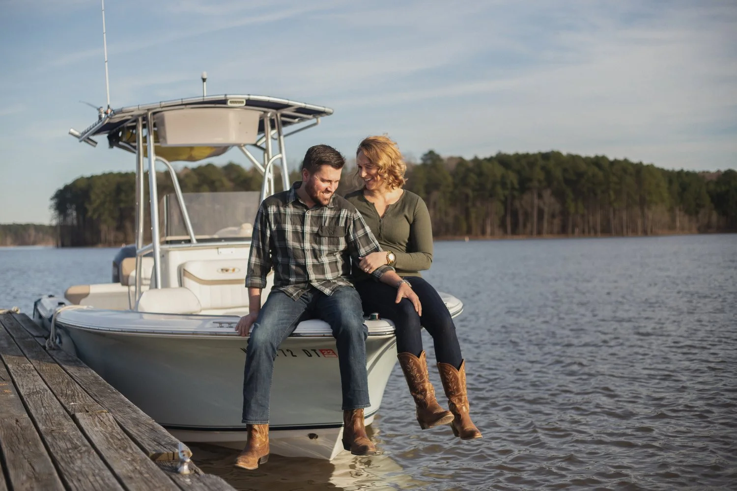A couple sits on the front end of a small white boat next to a wooden dock. The lake is seen around them and pine trees are across the water in the far distance behind them. He has on jeans, boots and a flannel type blue checked shirt. His left hand 