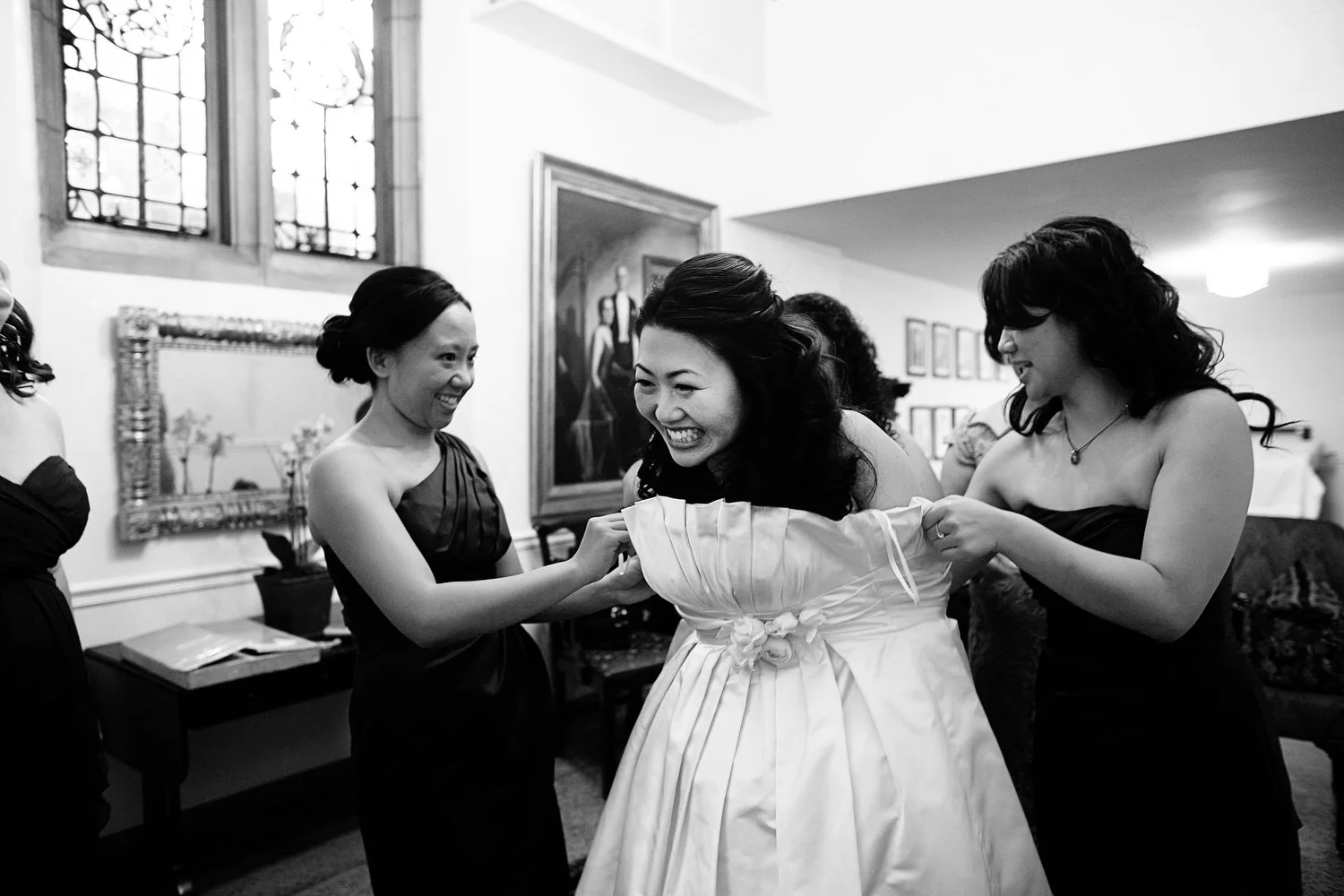 Black and white photo of a bride in a wedding dress smiling and being helped into her dress by her friends in a sitting room.