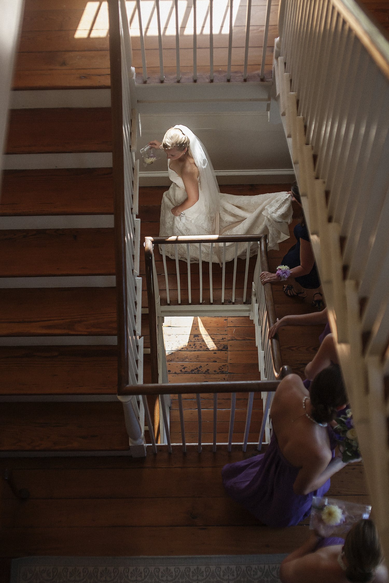 A bride in a white wedding dress and veil walking down a twisty wooden staircase. She is framed by steps she's already gone down in the foreground and appears small in between the railings in the distance.