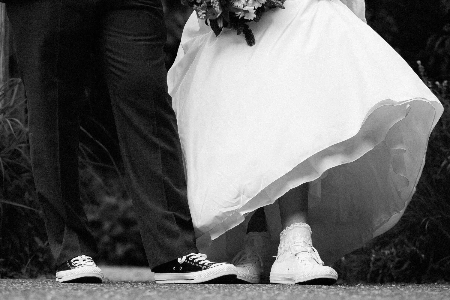 Tight black and white image taken from a low angle of a couple's feet. The male has on black dress pants and black converse shoes. The email has on a white wedding dress and is wearing white tennis shoes.