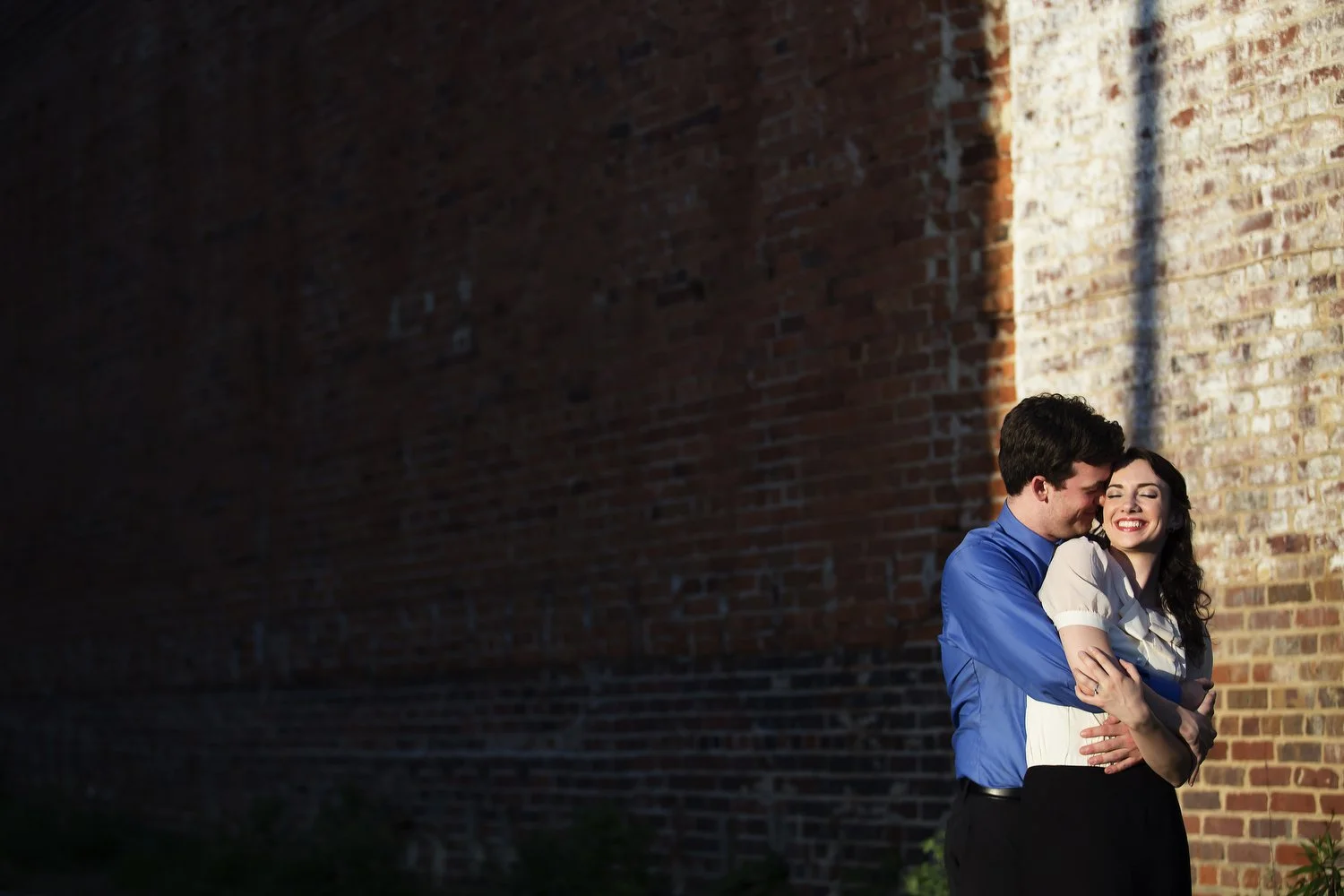 Loose photo taken at sunset. Around two-thirds of the image has a black shadow from a nearby building. A couple is seen in the far right section in the light near a brick wall. He has on a blue dress shirt and black pants and has his arms around her.