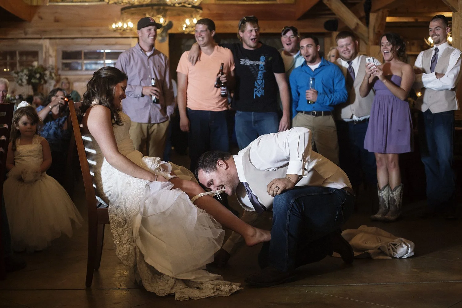 A bride with brown hair styled down in a white wedding dress sits in a chair as her groom, in a tan vest with a white dress shirt and jeans, kneels and removes her white garter with his teeth. Guests stand behind them and cheer and watch.