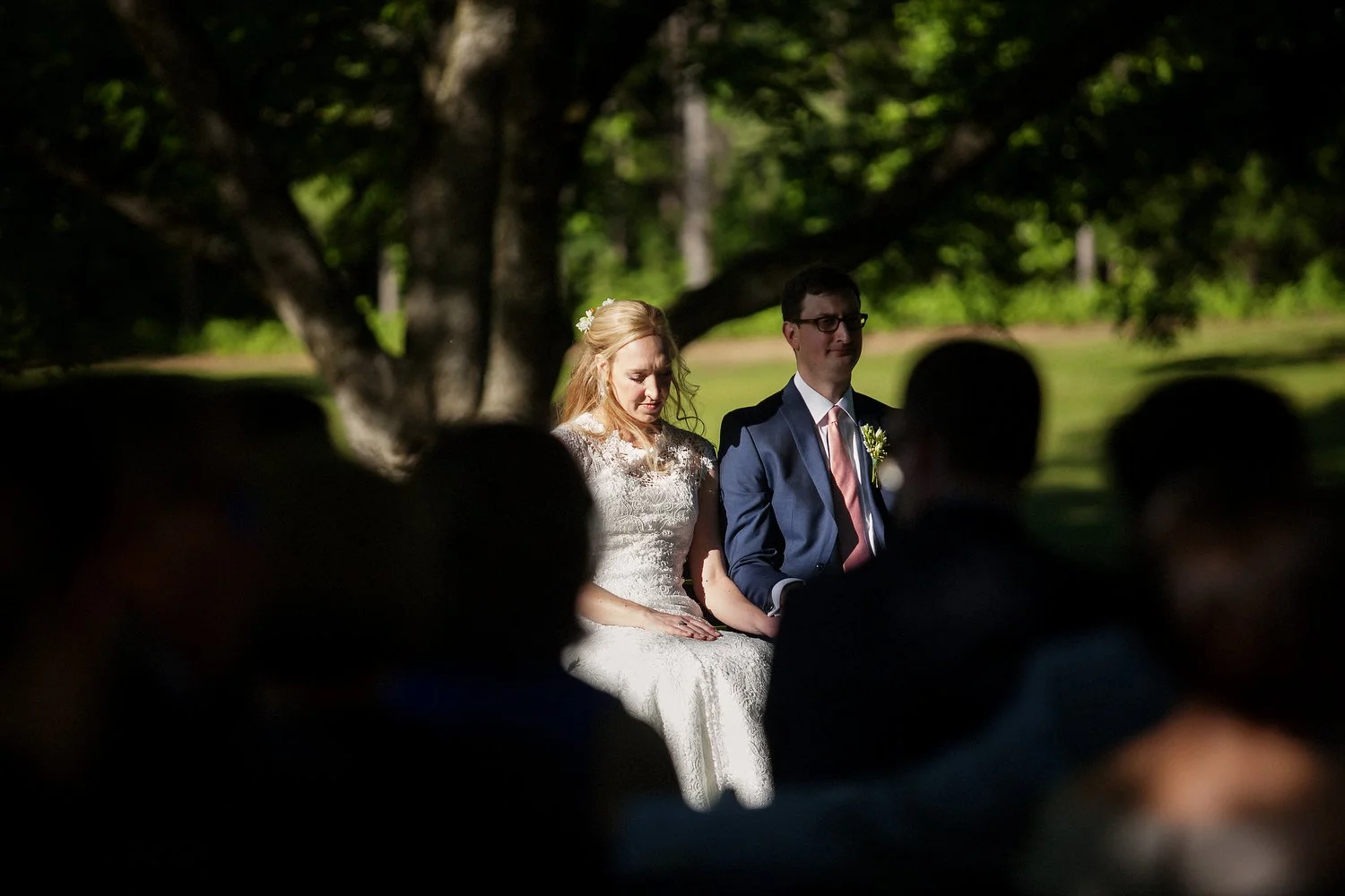 A bride and groom sitting outdoors under a large tree during their wedding ceremony, with guests silhouetted in the foreground.