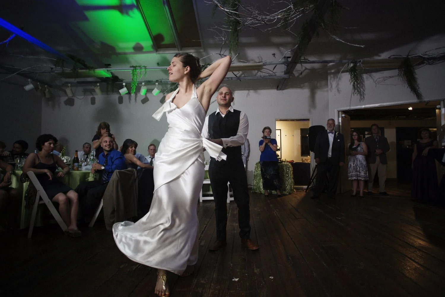 People sit at tables in the distance as a bride in a simple silk dress makes a move with her arm bent behind her head on the dance floor. There are green lights on the ceiling. A man in a black vest and dress pants and dress shirt stands in the dista