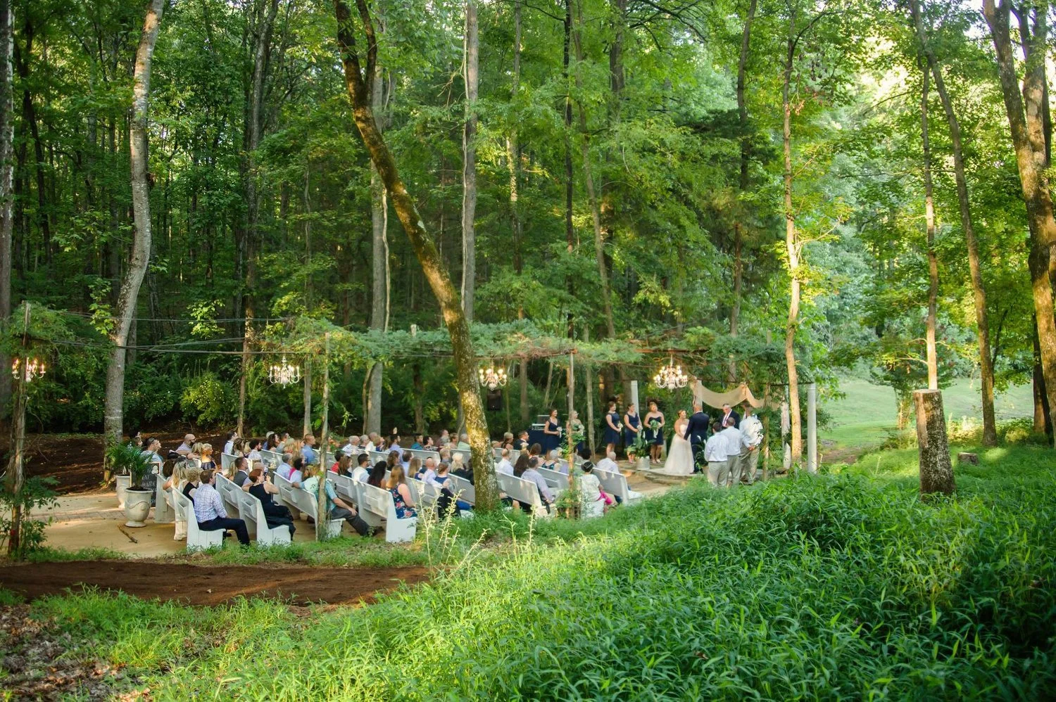 Outdoor wedding ceremony in a lush green forest with guests seated on white benches and bridesmaids standing with the bride and groom at the altar decorated with chandeliers and greenery.