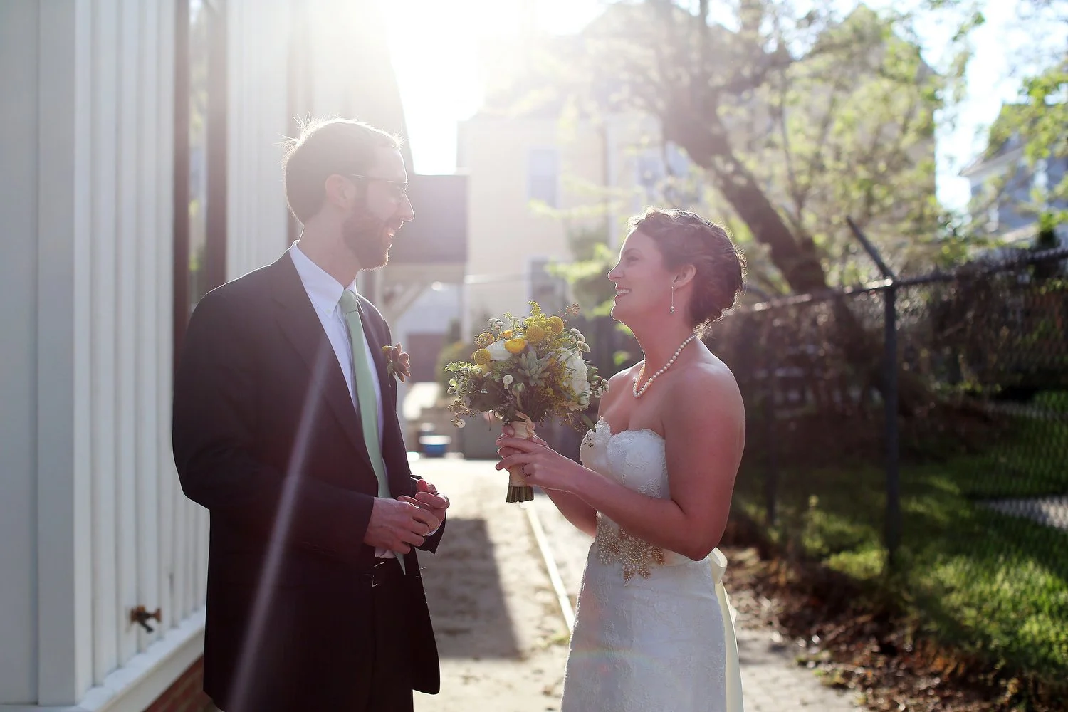A bride and groom share a moment outdoors on their wedding day. The bride is holding a bouquet of yellow and white flowers, and they are smiling at each other against a sunny, tree-lined background. He has a suit and glasses. She has a strapless whit