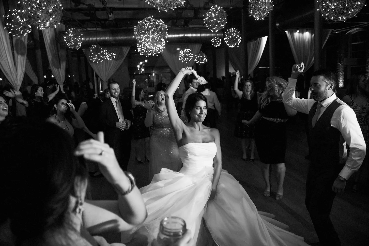 An elegant bride is in the center of a group of people on the dance floor. She clutches her strapless white wedding dress with her left hand as her right hand is arched up over her head as someone prepares to twirl her. A man with a vest and dress pa