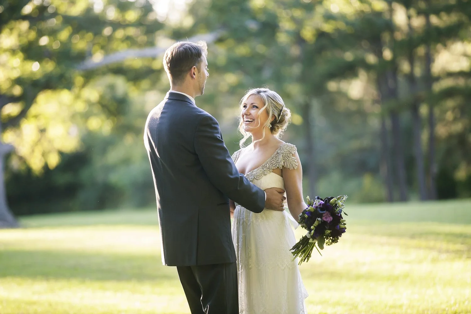 A bride and groom standing in a sunlit outdoor area, holding hands and looking at each other, with trees in the background. She is wearing a vintage dress with beaded details. He is in a suit.