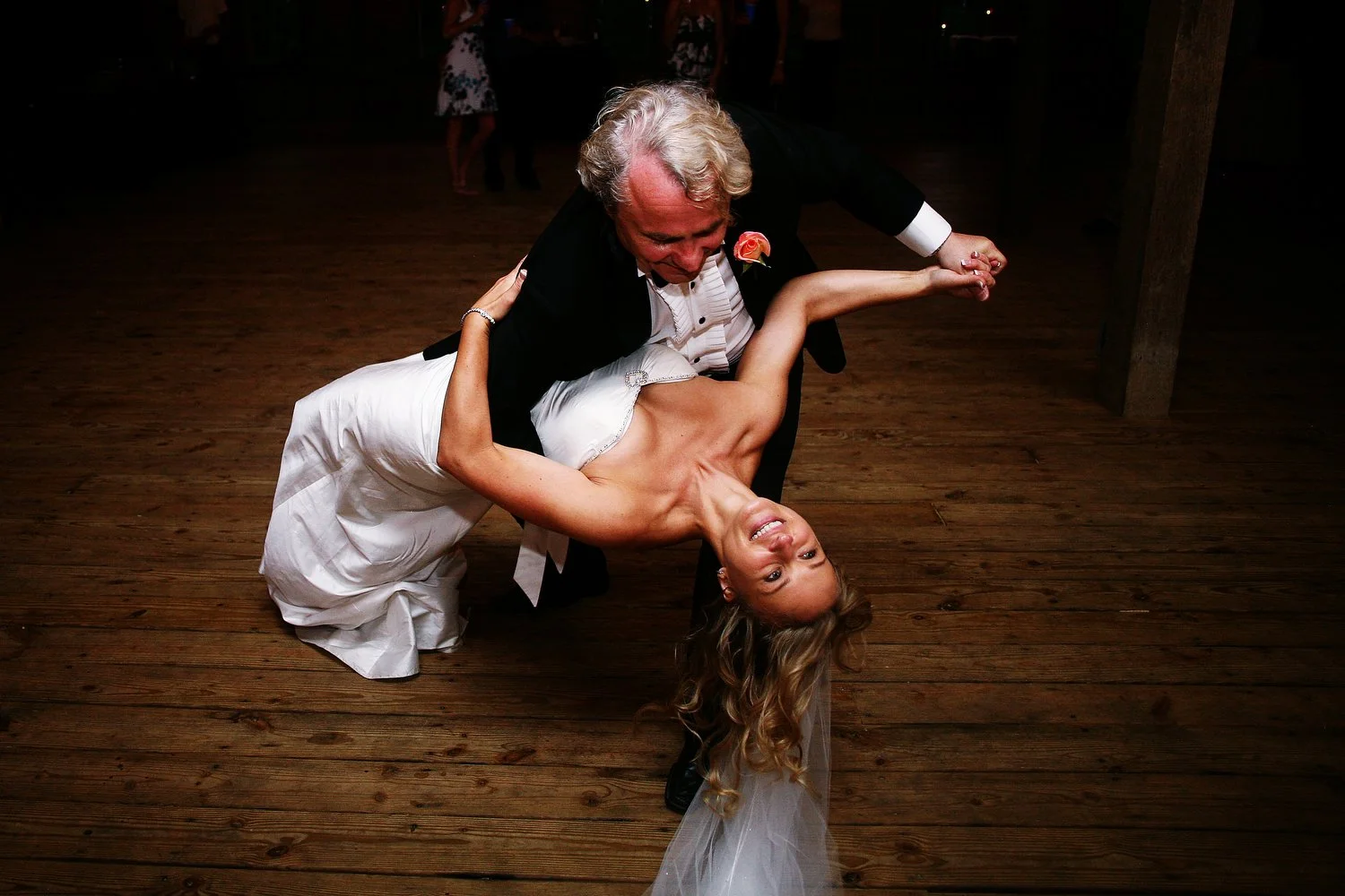 A bride in a strapless white wedding dress and a veil is dipped down toward the dance floor by an older man in a black tuxedo. He has grey hair and is perhaps her father. Her hair is blond and styled in loose curls.