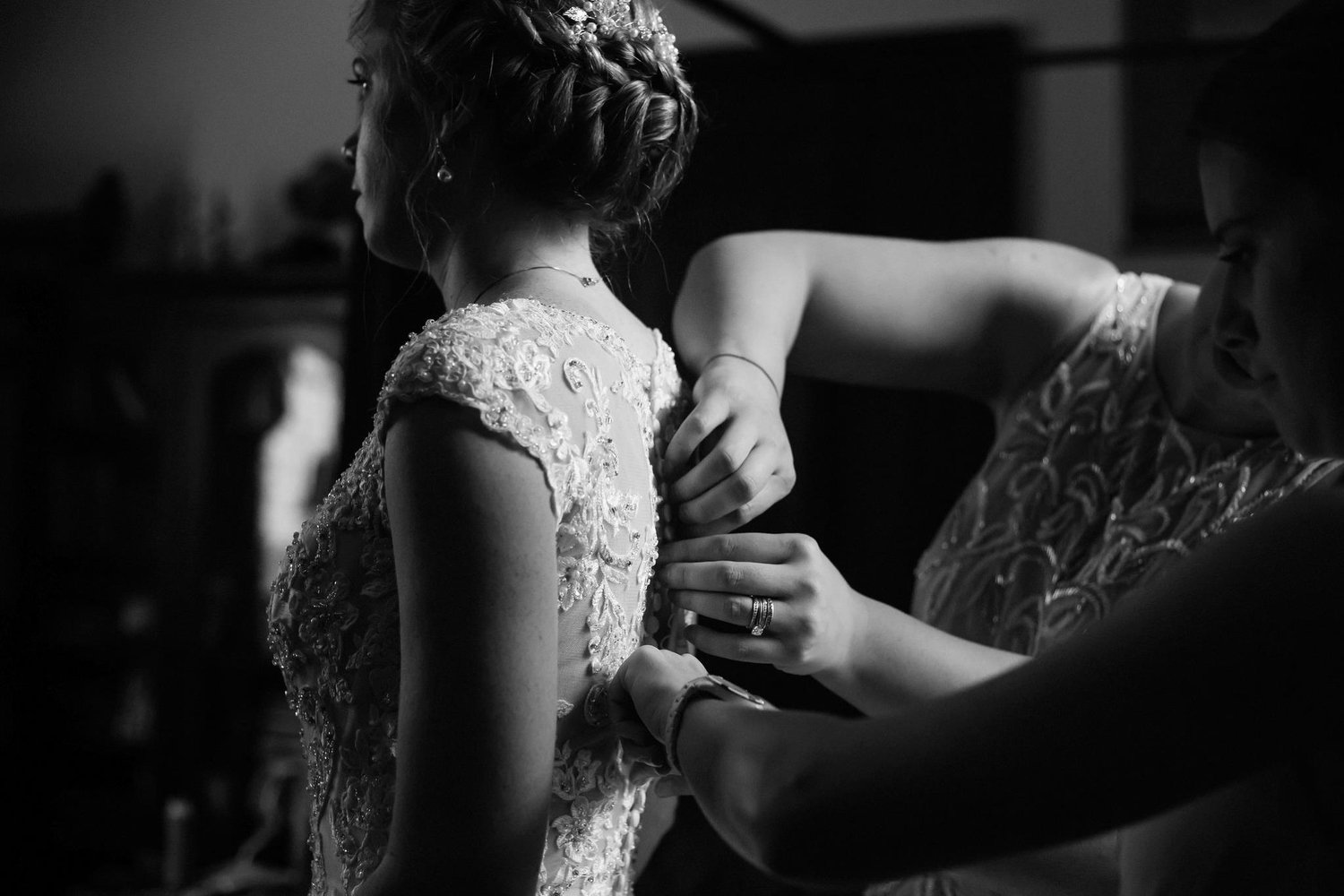 Black and white photo of a woman helping a bride get ready for her wedding by buttoning the back of her ornate lacy wedding dress.