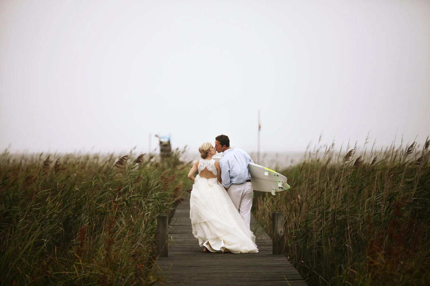 A bride and groom share a kiss on a wooden boardwalk surrounded by tall grass. The groom clutches a surfboard under his right arm. The view is from behind the couple as they walk to the water in the beach setting.