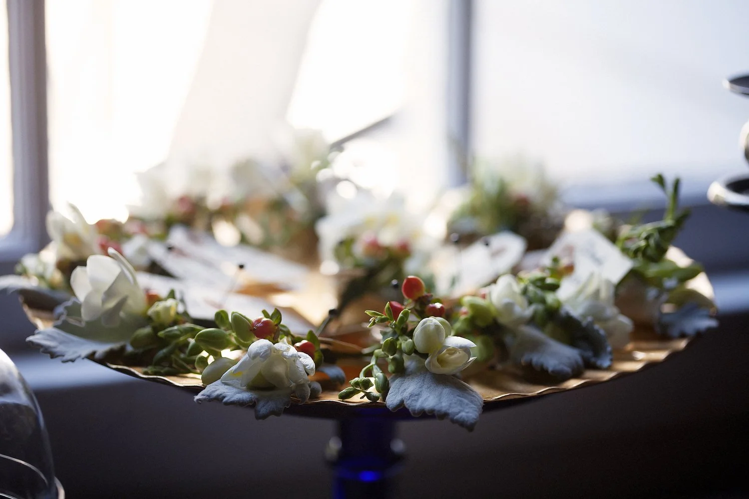 Several boutonnieres with small white and orange flowers are arranged around the edges of a circular decorative gold tray sitting in window light. 