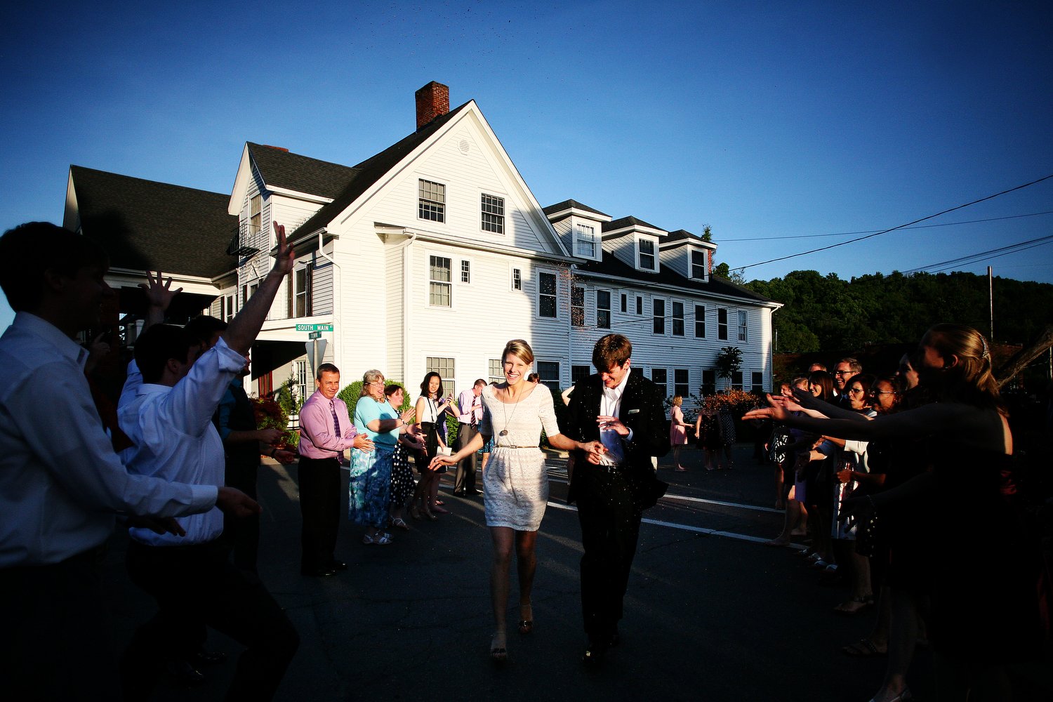 A group of people celebrating on a street in front of a large white house during the bride and groom's wedding reception exit in the late afternoon.
