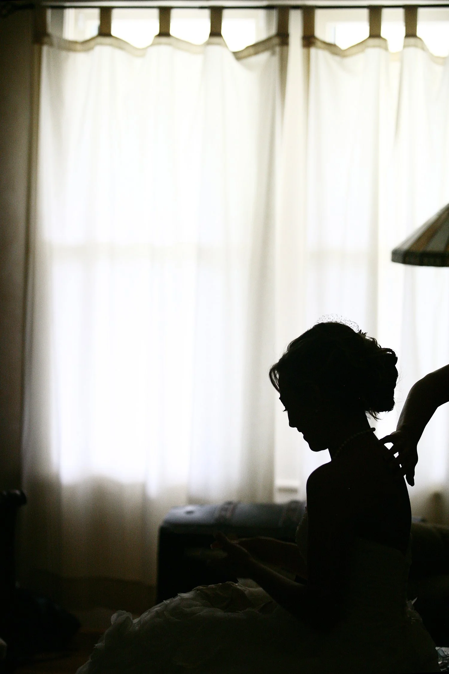 Silhouette of a woman in a white frilly dress and pearl necklace sitting in front of a lit window with curtains, with a person touching her back.